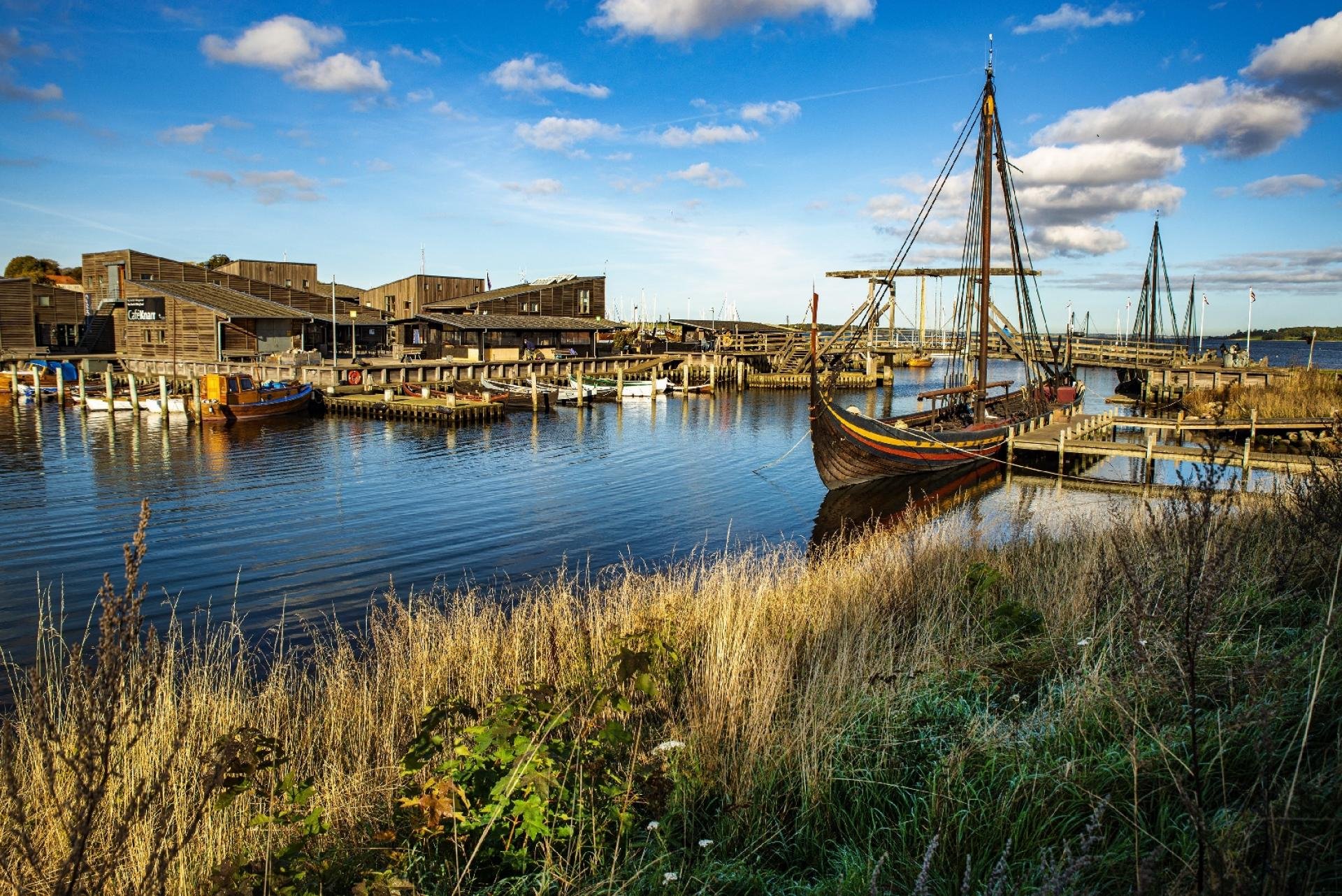 En havn med både moderne og gamle træskibe, omgivet af træbygninger, under en blå himmel med nogle skyer.