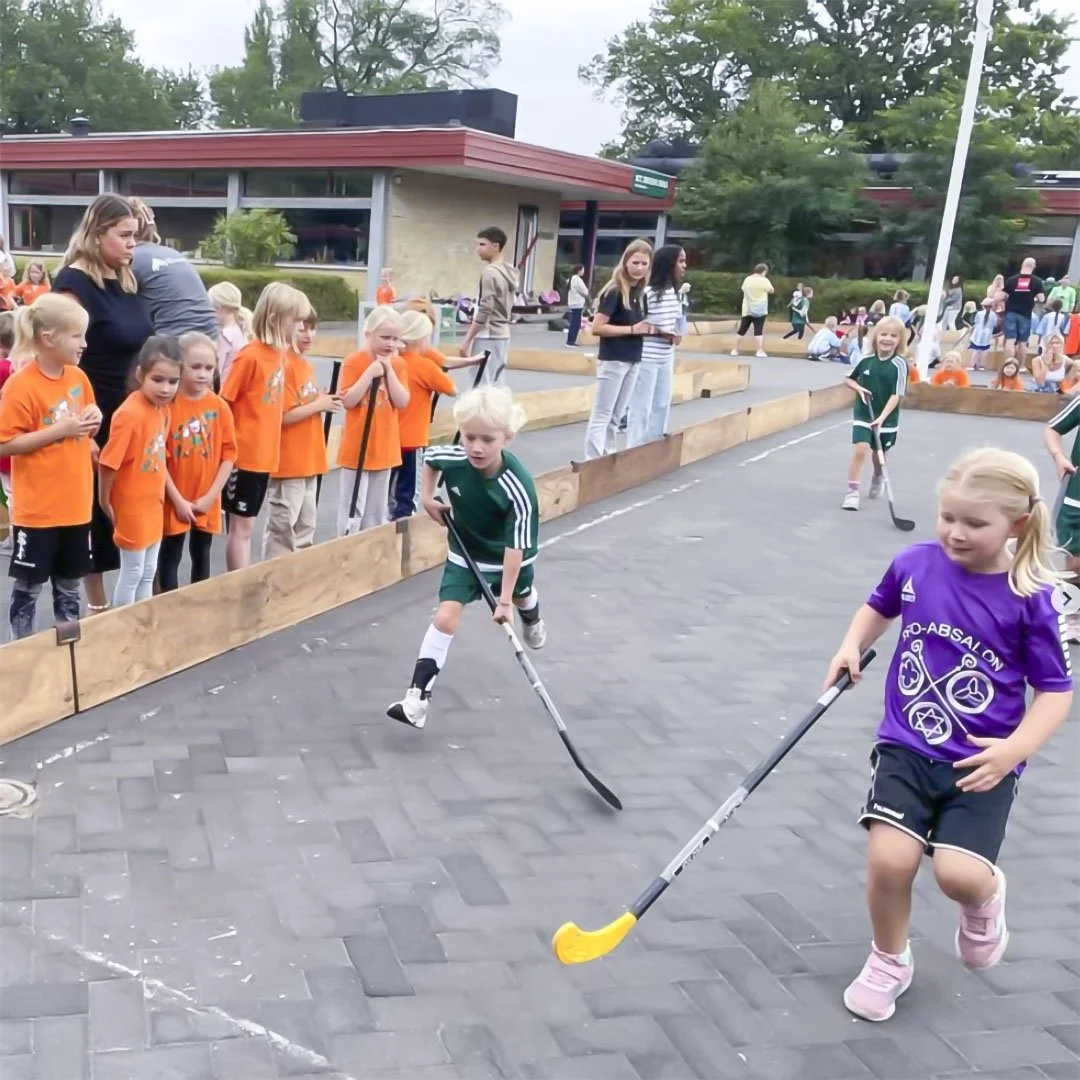 Børn spiller hockey på udendørs bane med tilskuergruppe og skolebygning i baggrunden.