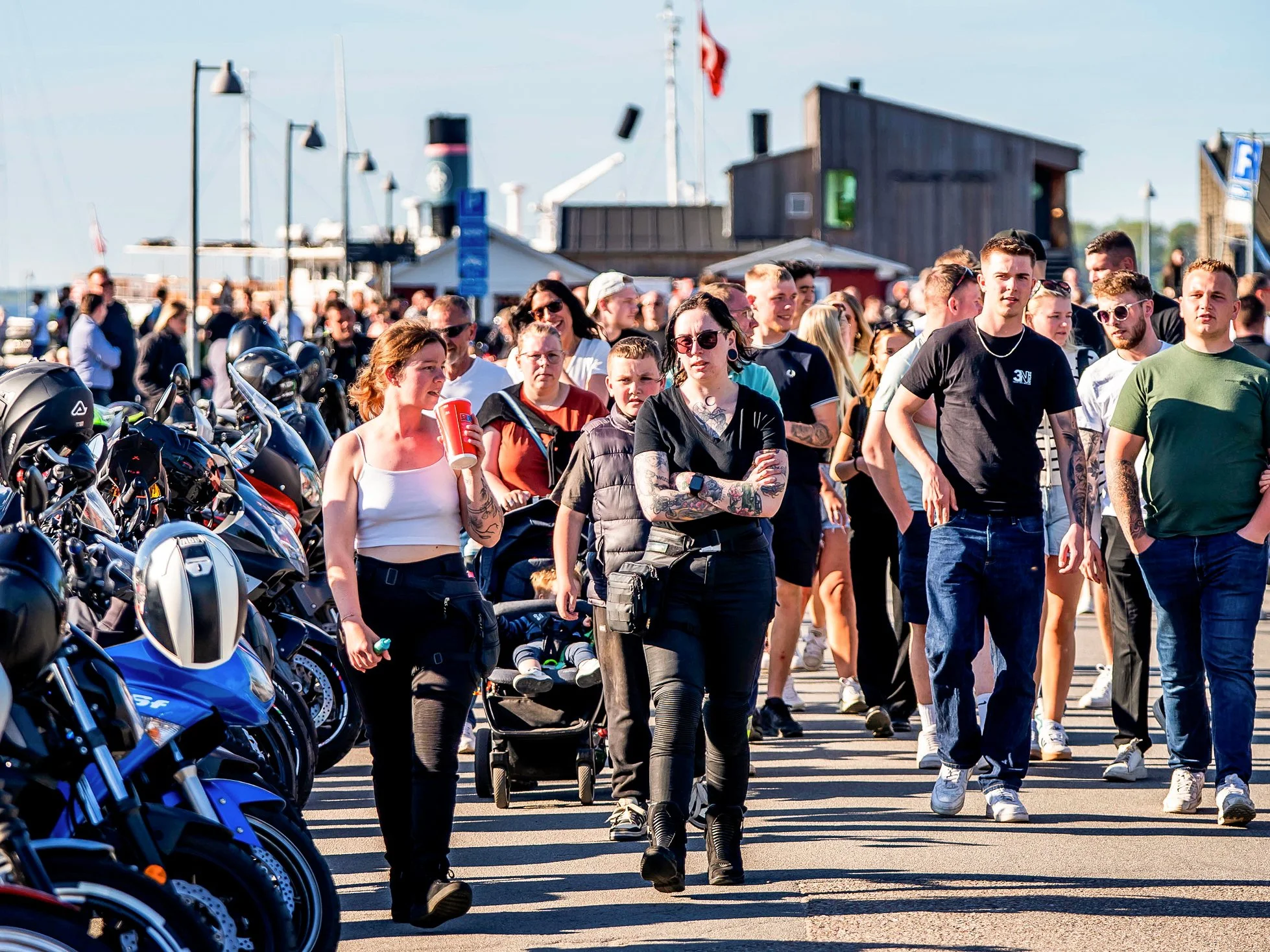 Mennesker går langs en parkingordning med motorcykler, mens baggrunden viser en havn med en stor historisk skib og en bygning, under en klar blå himmel.
