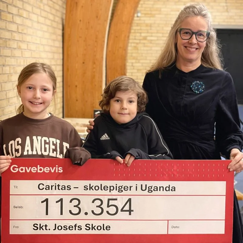 En kvinde og to børn holder et stort gavekort til Caritas for skolepiger i Uganda. Kvinden har blondt hår og briller, børnene er en pige med lyst hår og en dreng med brunt krøllet hår.