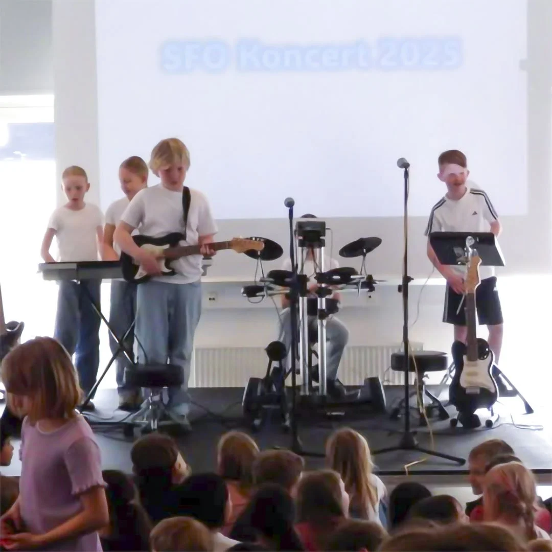 Børn optræder på en scene med musikudstyr foran et publikum, mens der vises et slideshow i baggrunden med teksten "SEO Koncert 2023".