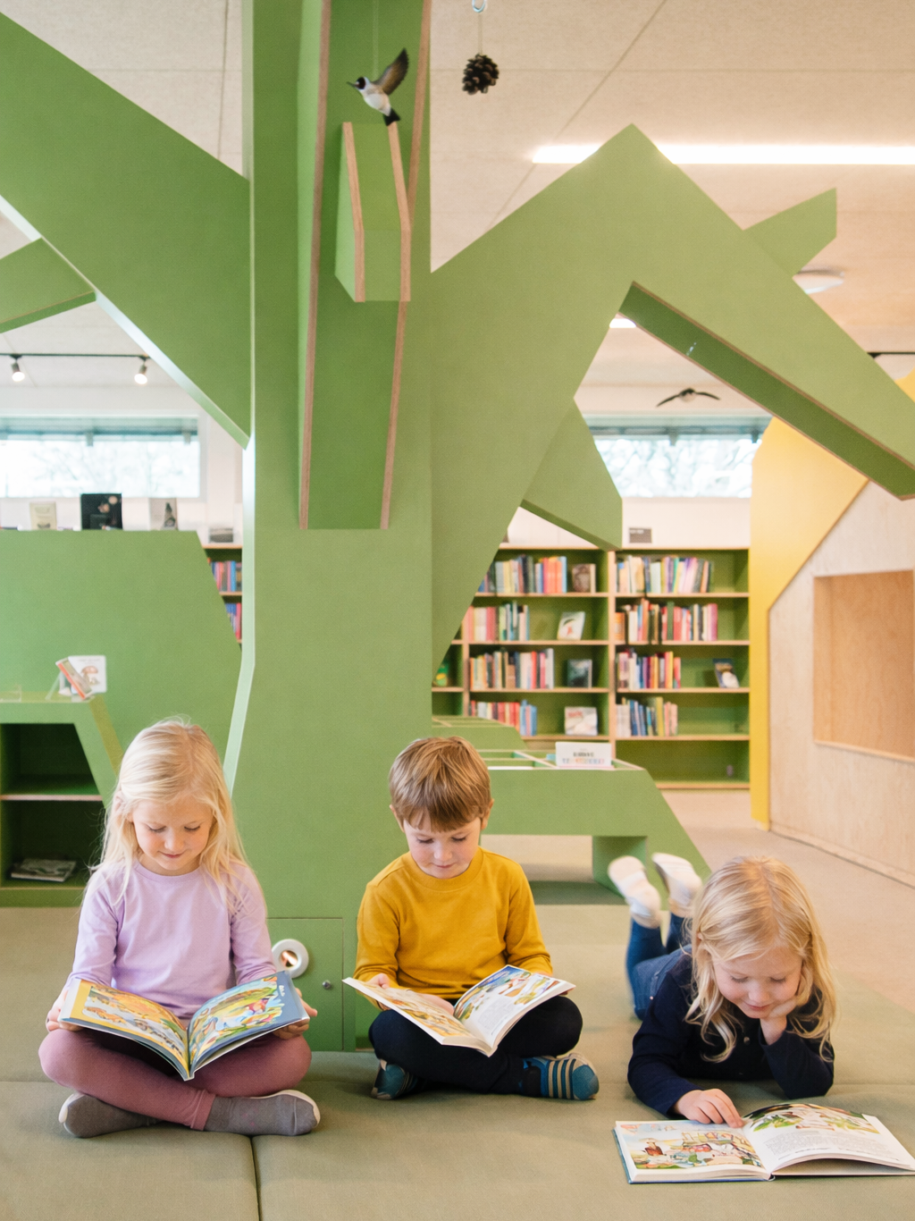 Tre børn sidder på gulvet og læser bøger i et bibliotek med grøn og gul vægdekoration, der ligner et træ. Der er bøger på hylder i baggrunden.