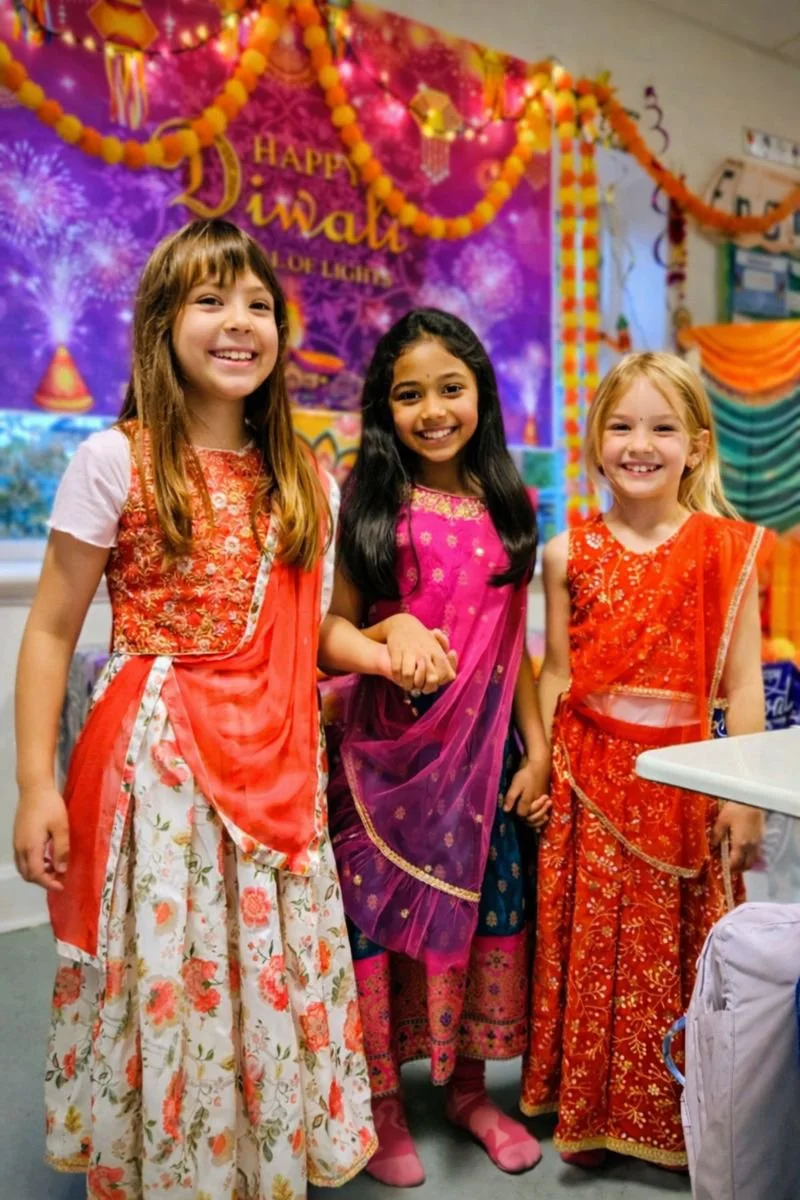 Tre piger står sammen og smiler iført farverige traditionalle indiske outfits julefest pynt i baggrunden