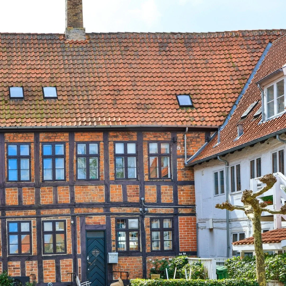 Oldt hus med brændte teglsten på taget og rødbrune murstensvægge, vinduer med små rammer, og en hvid bolig med hvide vinduer og en beskåret træ i haven.