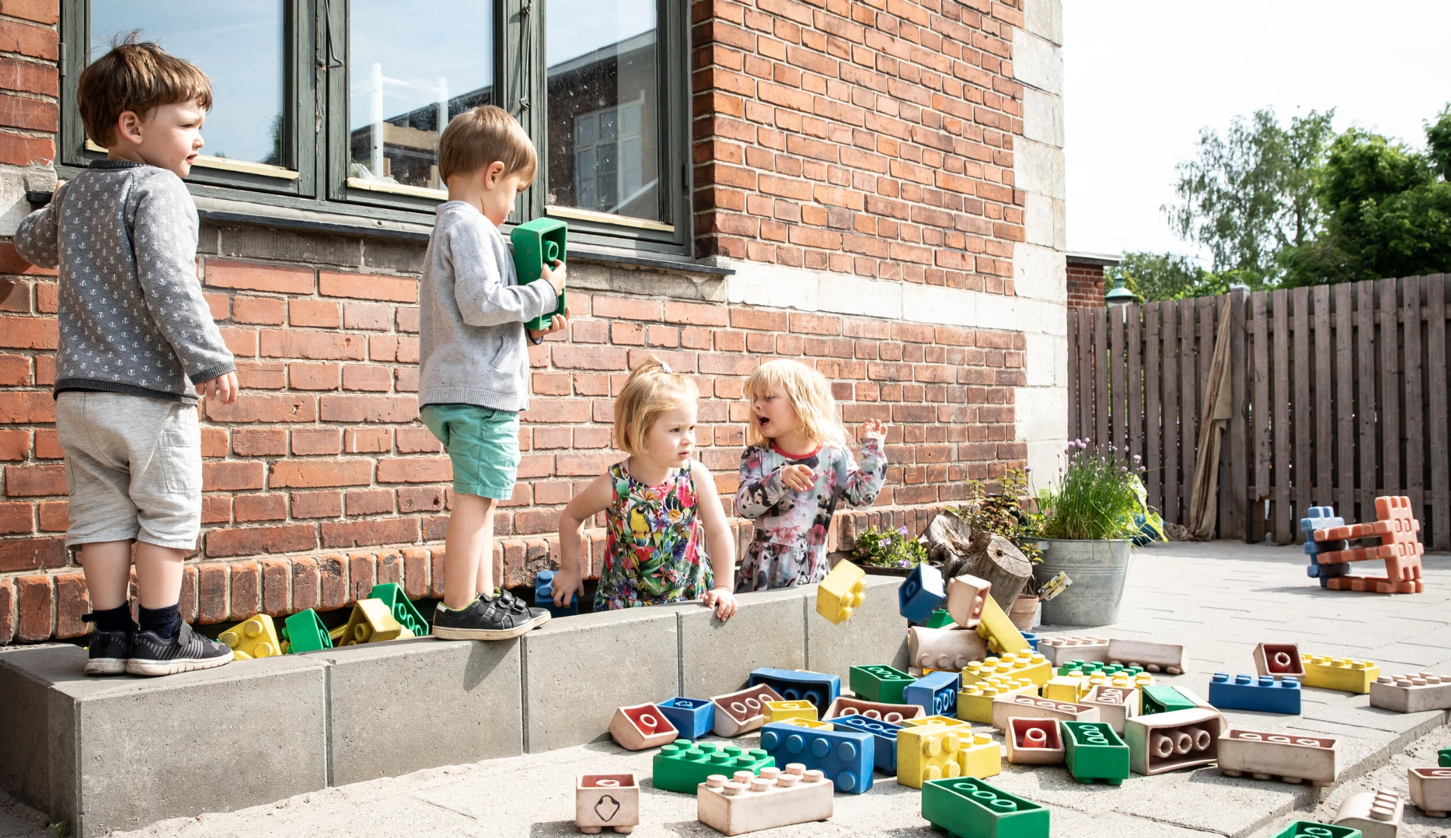 Fire børn leger udenfor, med legoklodser på en sand- og stenbelagt gård ved en rødbrun mur, virt i en udendørs pædagogisk playground.