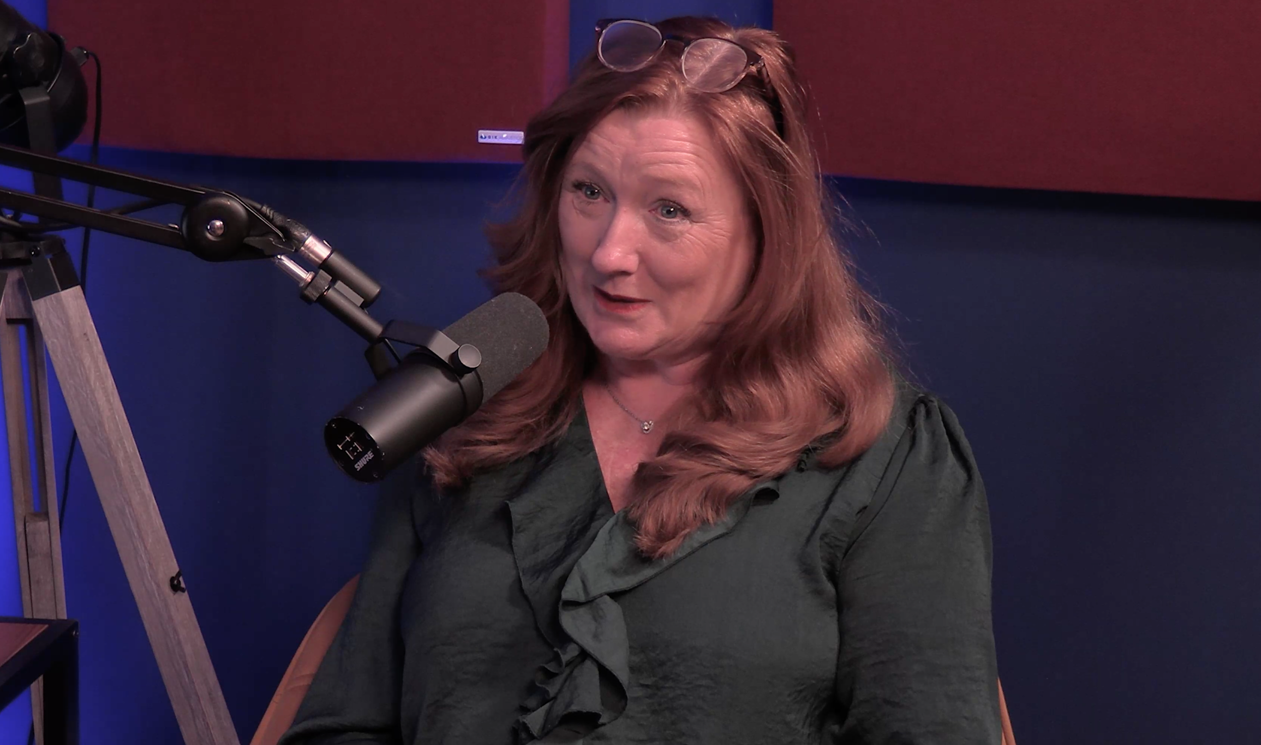 A woman with red hair and glasses resting on her head, sitting in front of a microphone, speaking or listening in a studio with a dark blue and red background.