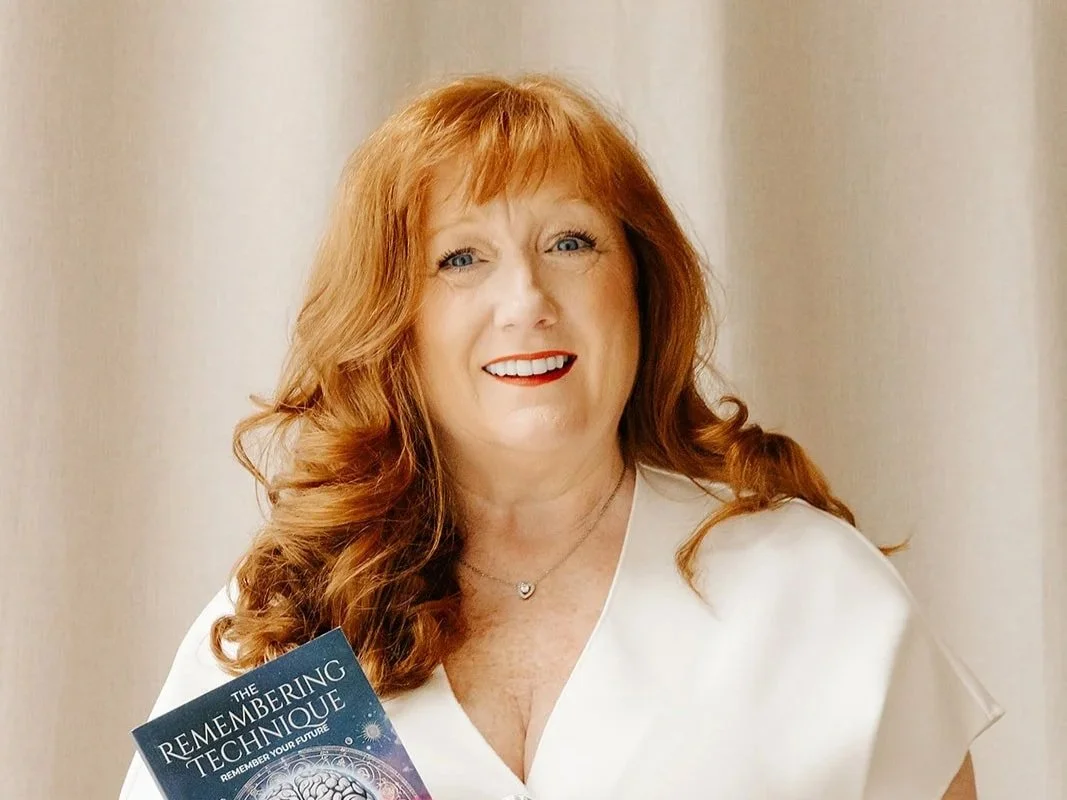 A woman with red hair and a white top holding a book titled 'The Remembering Technique' and smiling at the camera.