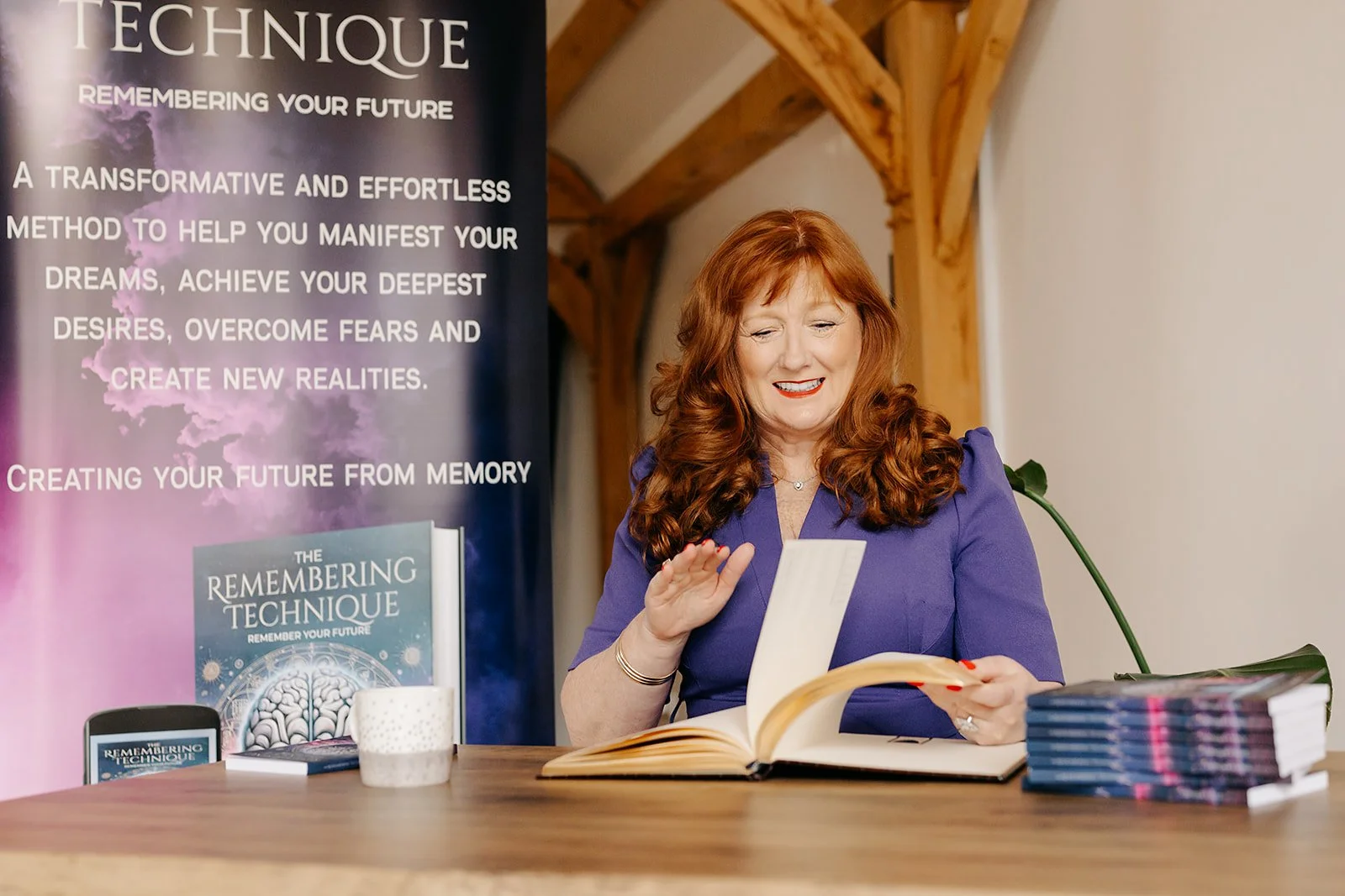 A woman with red hair, wearing a purple dress, sitting at a wooden table, smiling and looking at a book in her hands. Behind her is a banner that reads 'The Remembering Technique' and describes a method to manifest dreams and create new realities. The table has a stack of books, a white mug, and a smartphone displaying the same book cover.