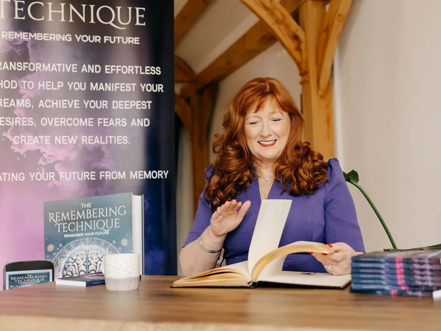 A woman with red hair is sitting at a table, smiling and looking at a book she is flipping through. There are stacks of books, a white mug, and a promotional banner for 'The Remembering Technique' behind her.