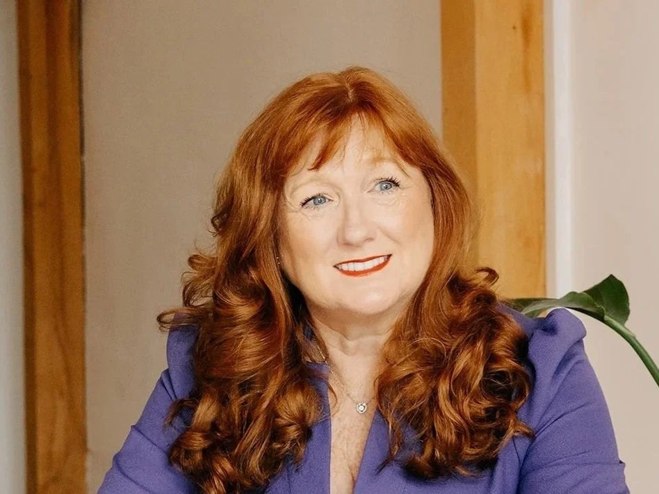 A woman with long red hair, blue eyes, and red lipstick, wearing a purple top, sitting indoors with a wooden wall and a green plant in the background.
