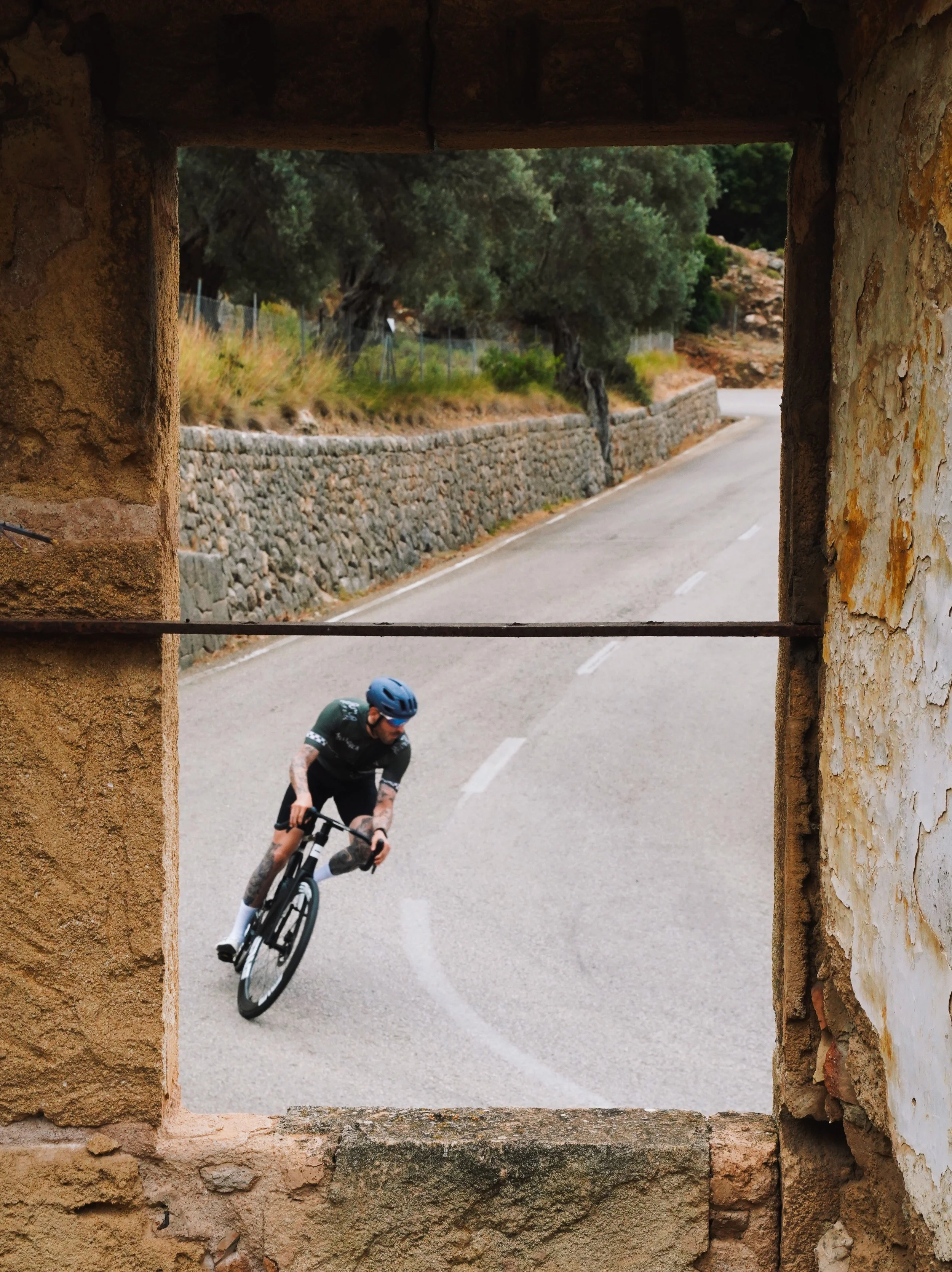 A cyclist wearing a helmet and black outfit riding a bicycle around a bend on a mountain road, seen through a stone window.