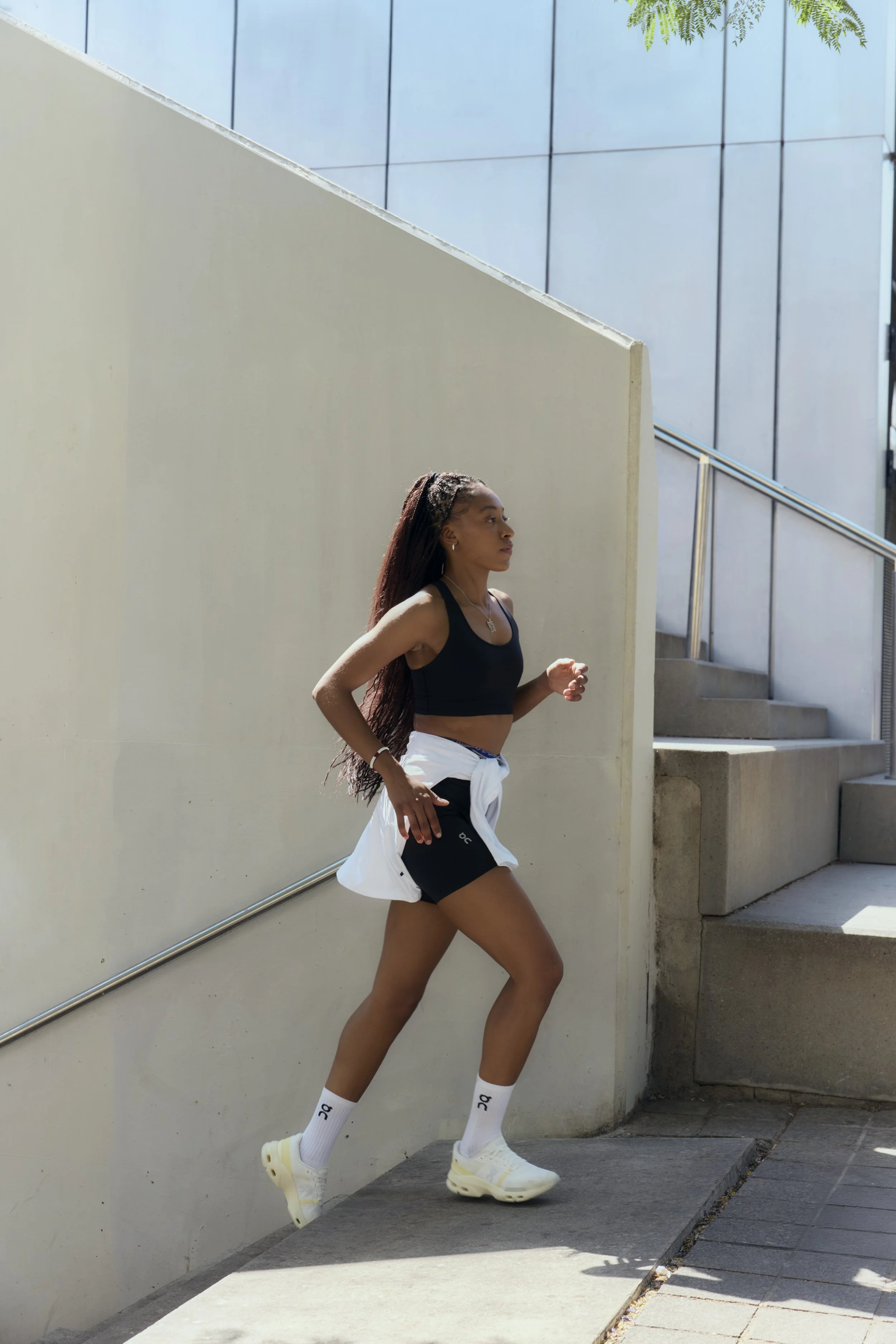 Young woman running outdoors in urban setting, wearing athletic shorts, tank top, white jacket tied around waist, white socks, and sneakers.