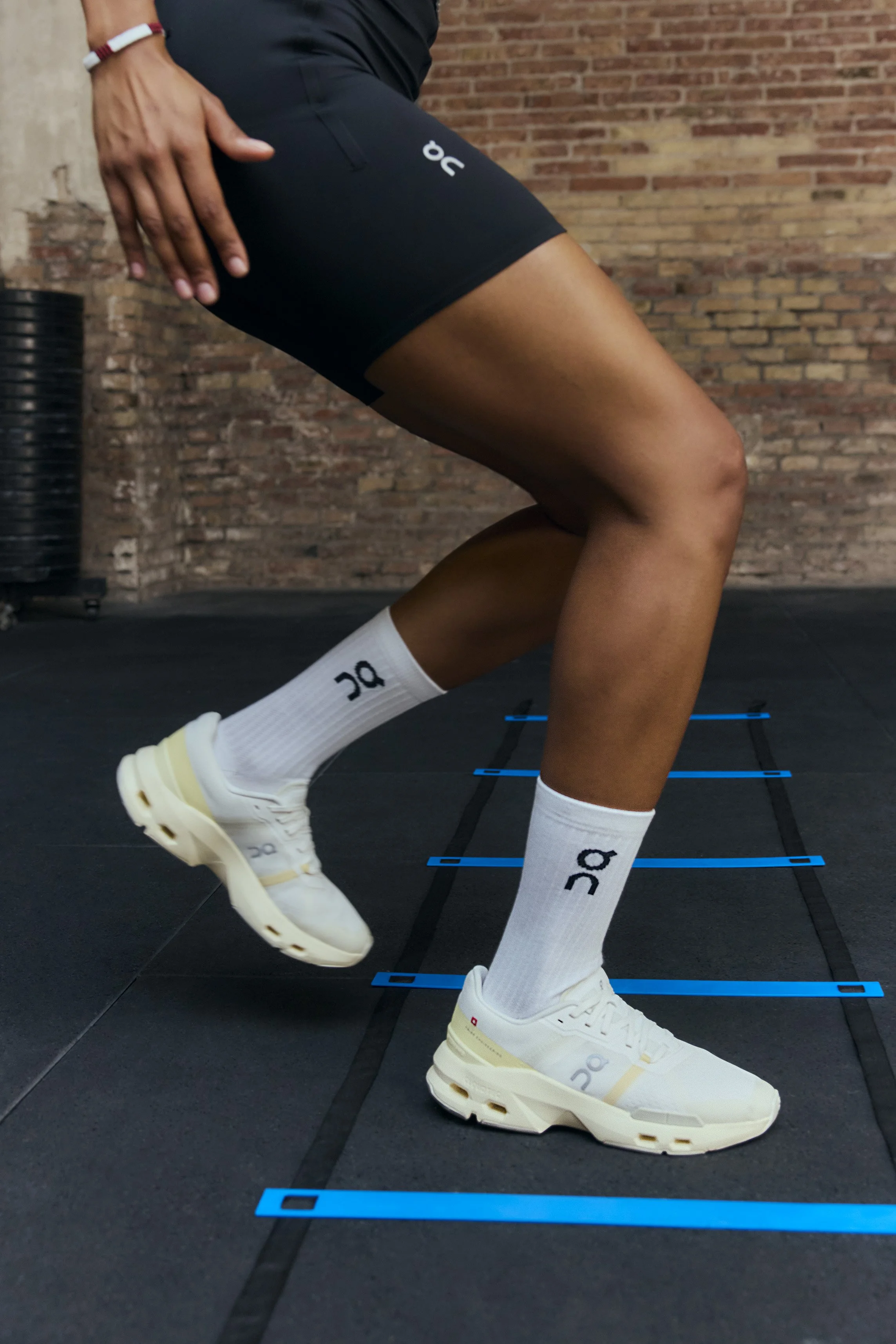Person wearing white athletic shoes, white socks with a logo, and black athletic shorts, performing an exercise on an agility ladder in a gym setting with brick wall in the background.