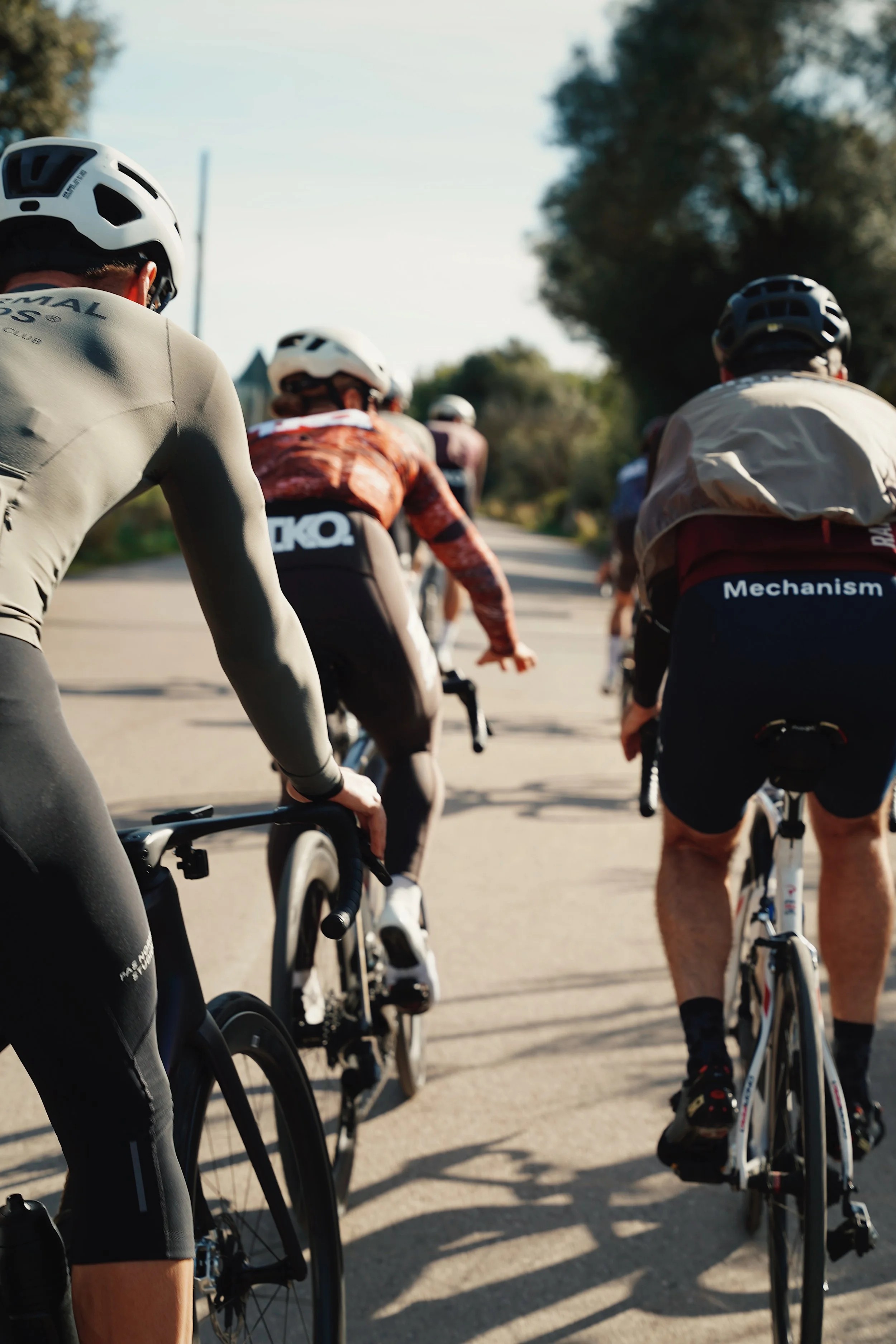 A group of people riding bicycles on a road, wearing helmets and athletic clothing, with trees in the background.