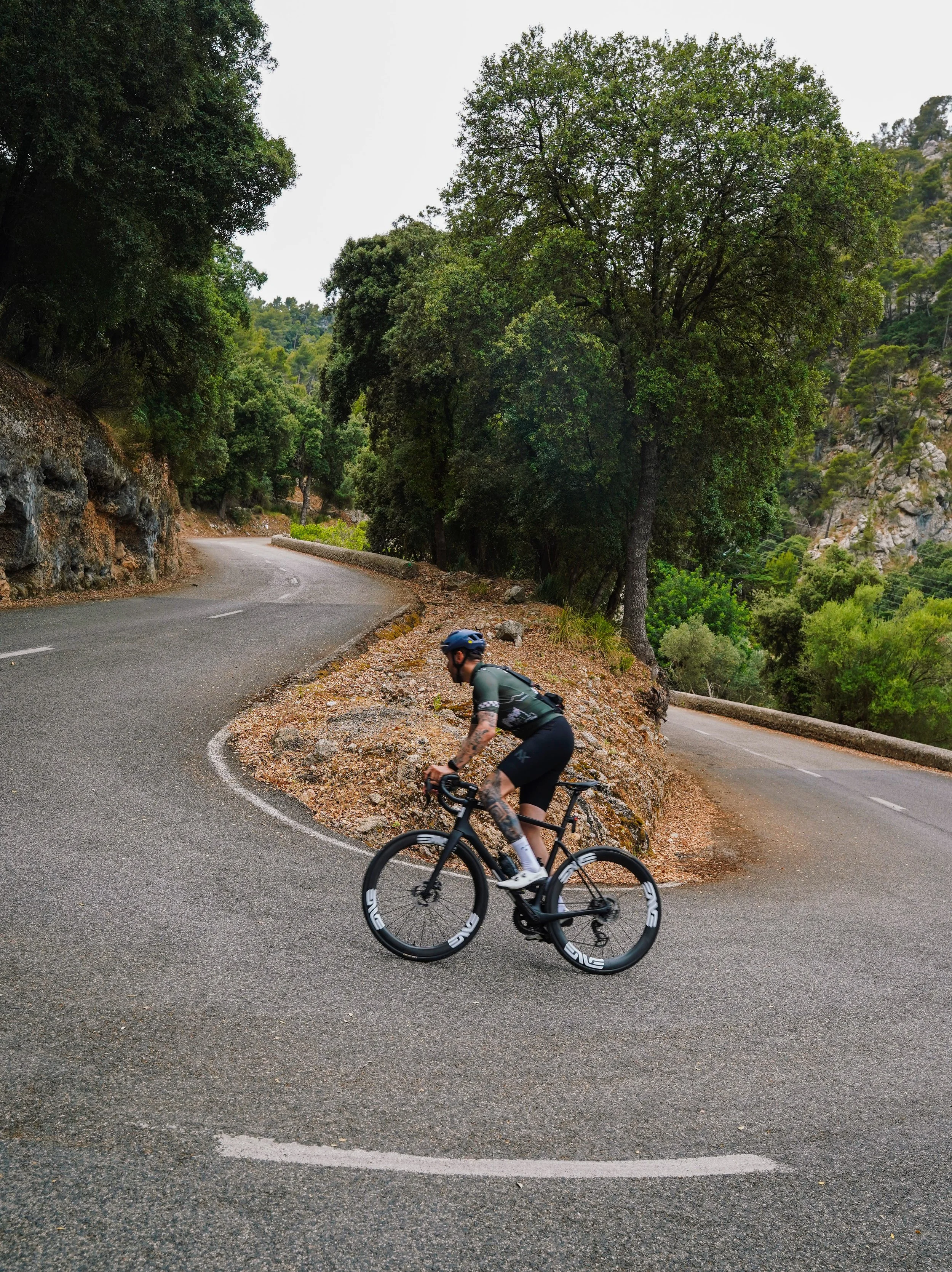 A cyclist wearing a helmet and sunglasses riding a black bike on a winding mountain road surrounded by trees and rocky terrain.