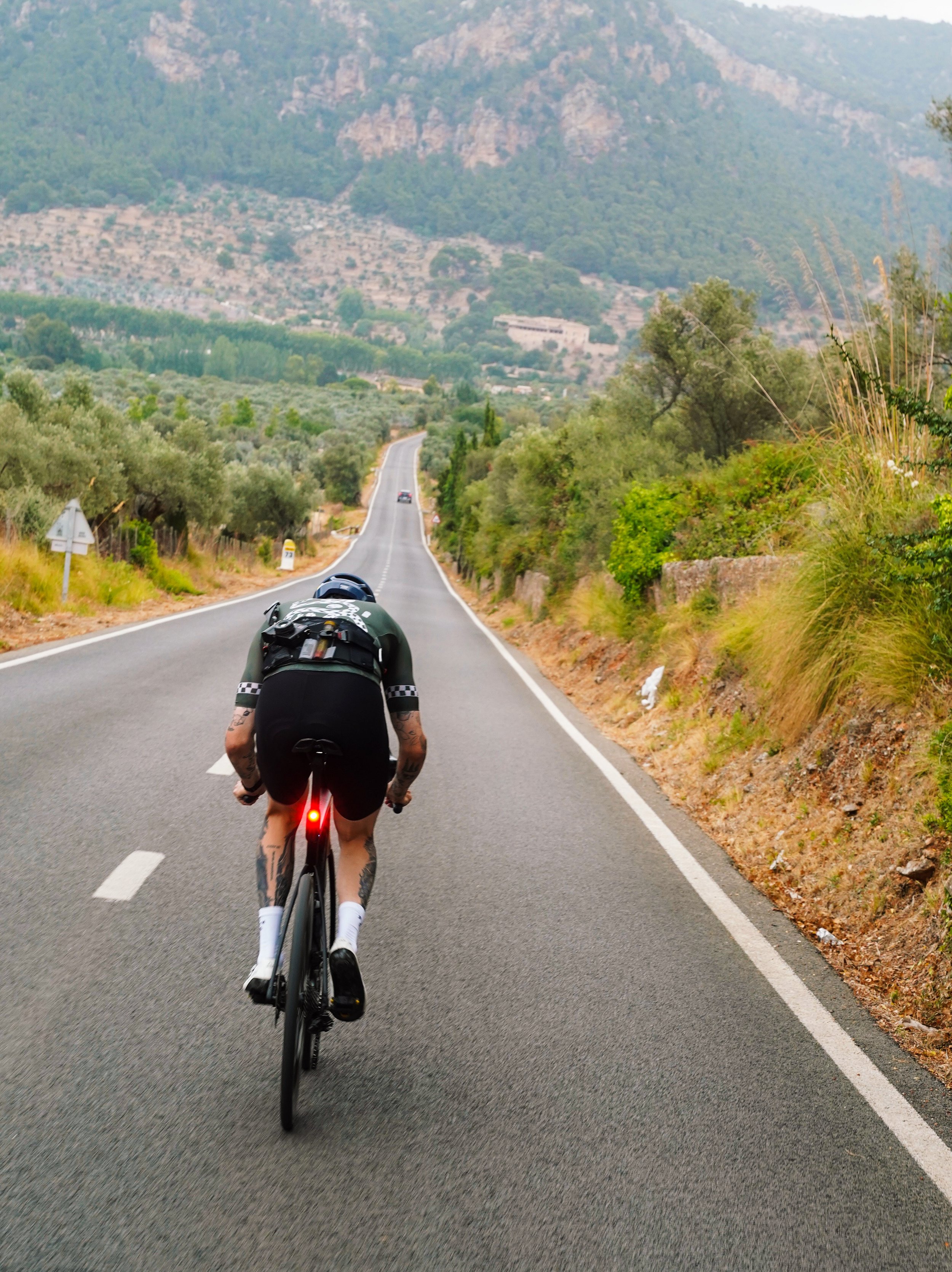 A cyclist riding on a winding rural road surrounded by trees and mountains in the background.