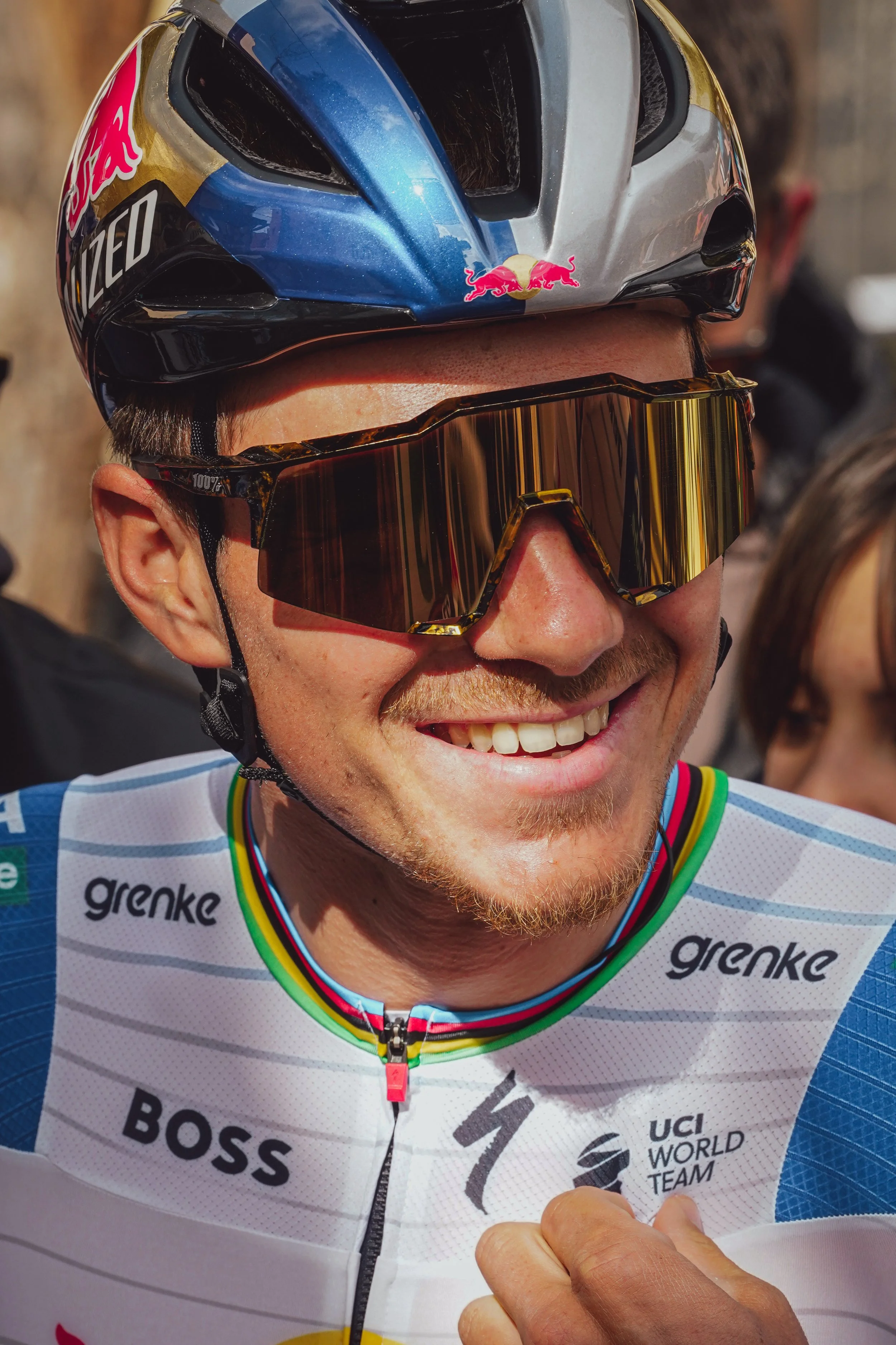 Close-up of a smiling male cyclist wearing a blue helmet, tinted sunglasses, and a white cycling jersey with sponsorship logos, outdoors during daytime.