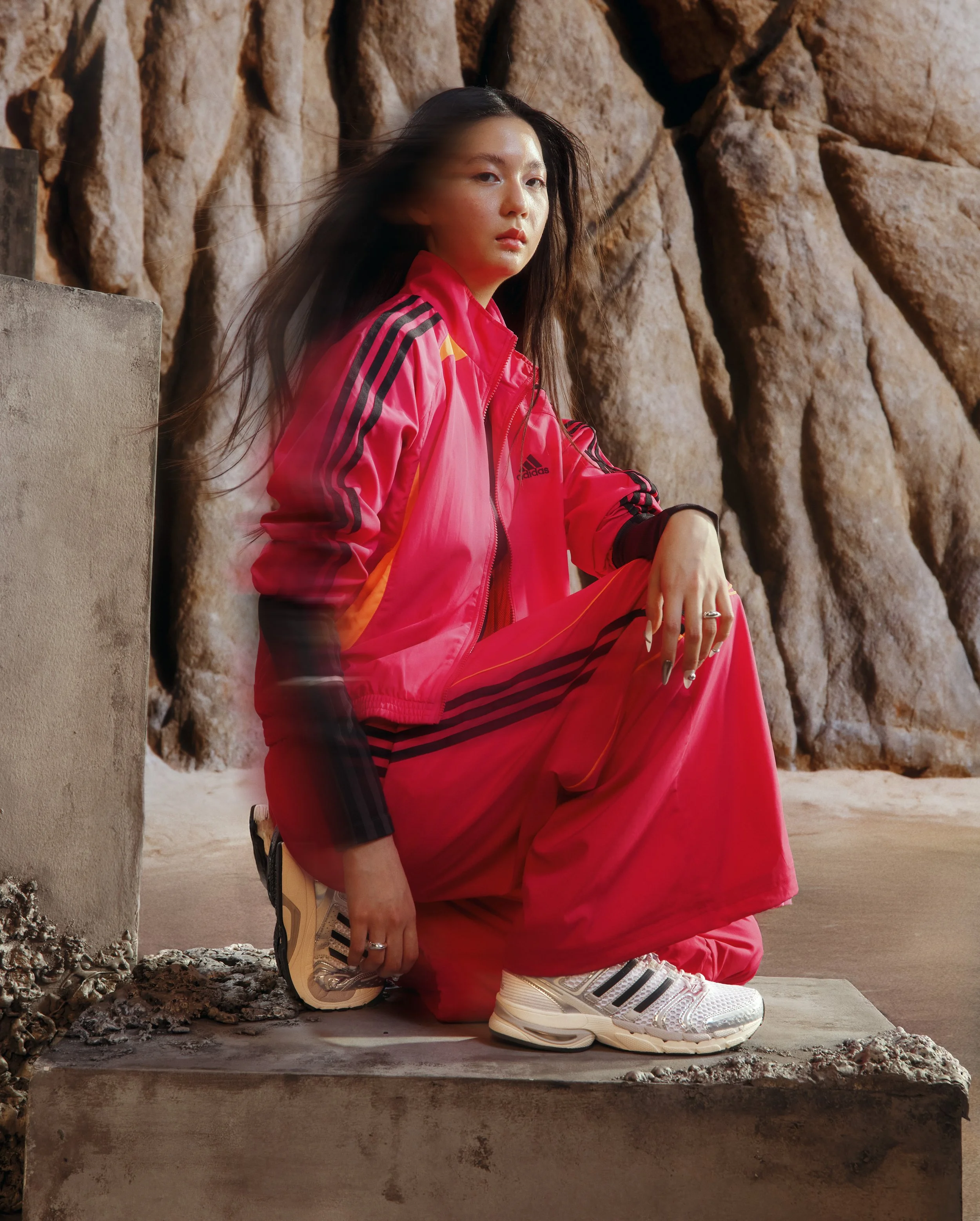 A woman dressed in a bright pink Adidas tracksuit squatting on a concrete platform near rocky terrain.