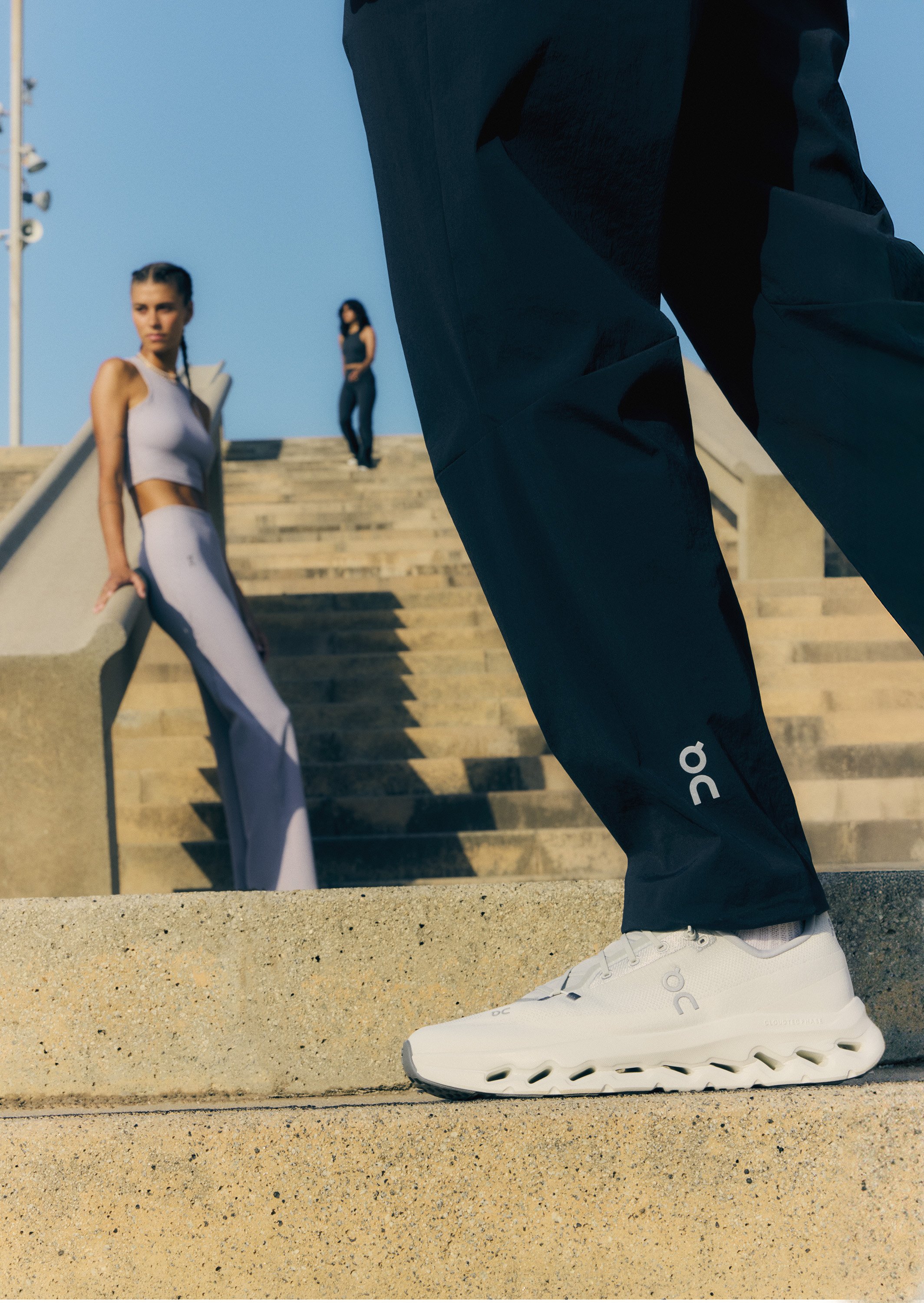 Close-up of a person wearing white athletic shoes and black Nike On running pants, standing on concrete steps with two women in workout clothing in the background.