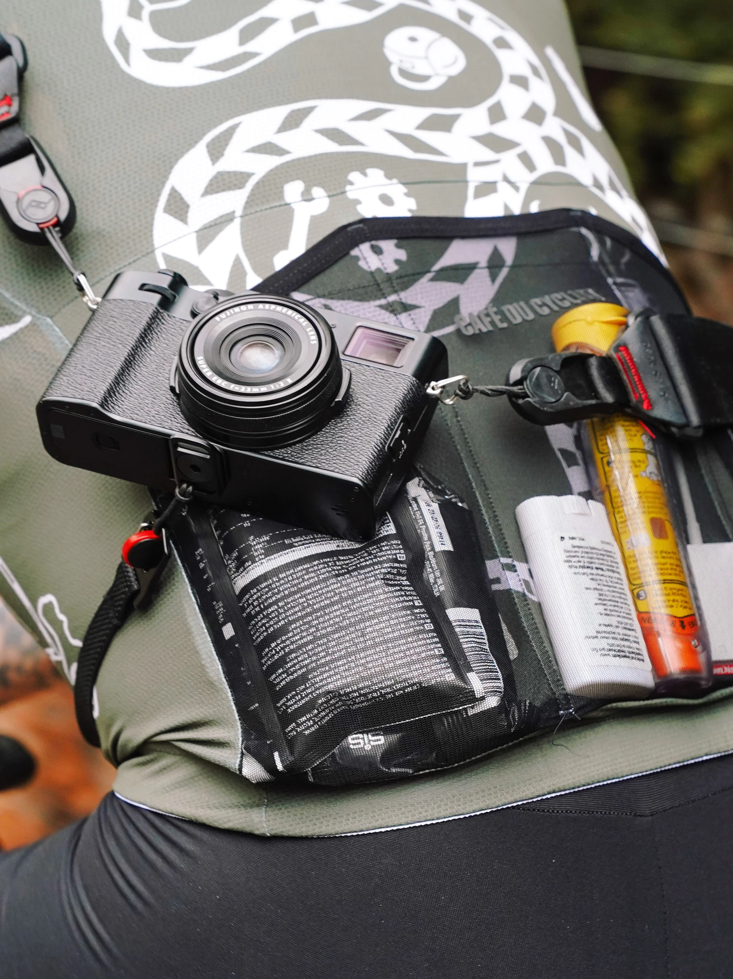 A black digital camera resting on a green and black backpack with multiple compartments, holding a yellow travel-sized spray, a white container, and an orange tube, on a person wearing black outdoor pants.