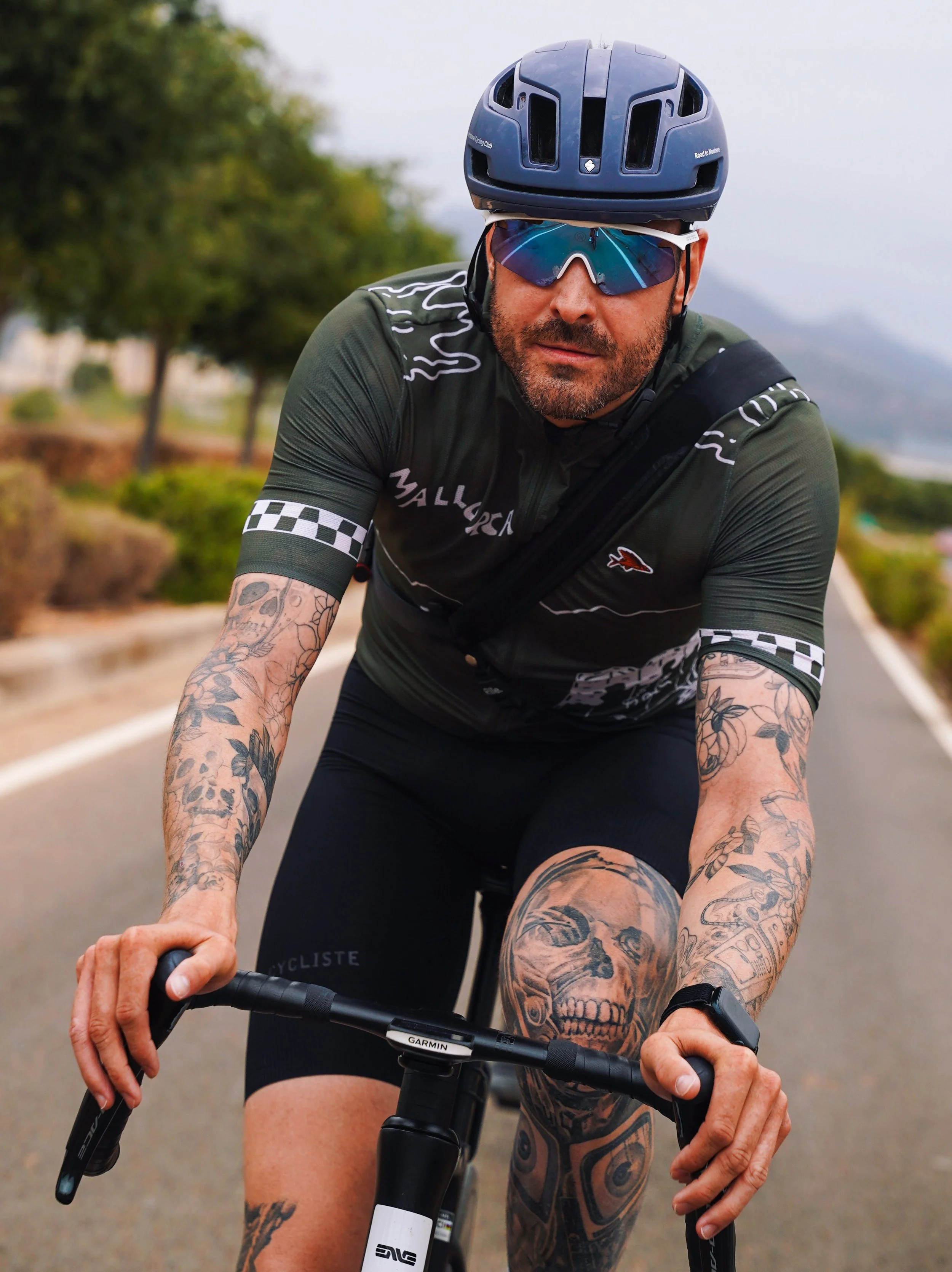 A man wearing a blue bike helmet, sunglasses, and a green cycling jersey is riding a bicycle on a paved trail. The man has tattoos on both arms and legs, and is focused on riding.