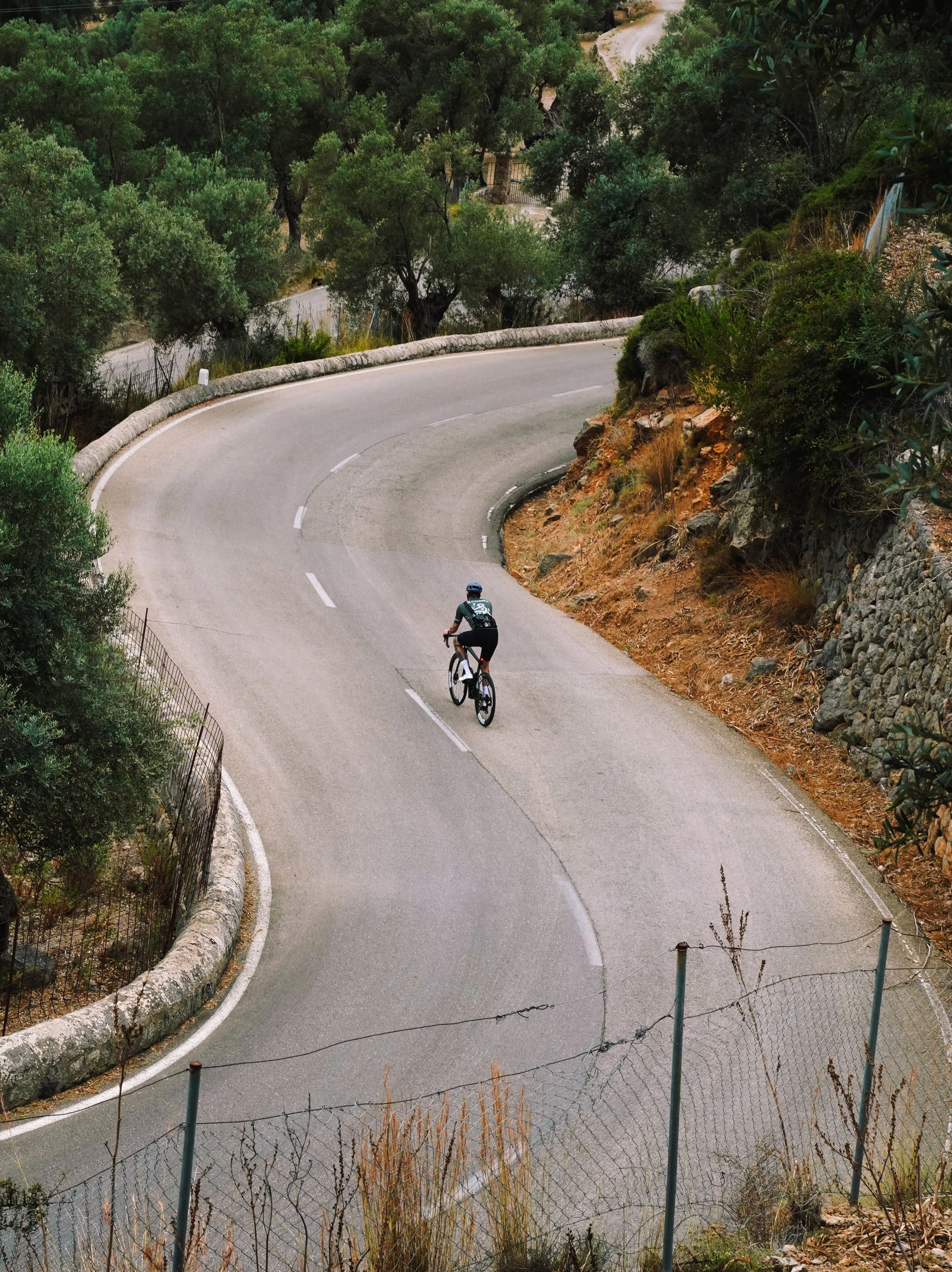 A person riding a bicycle on a winding mountain road surrounded by green trees and rocky terrain.