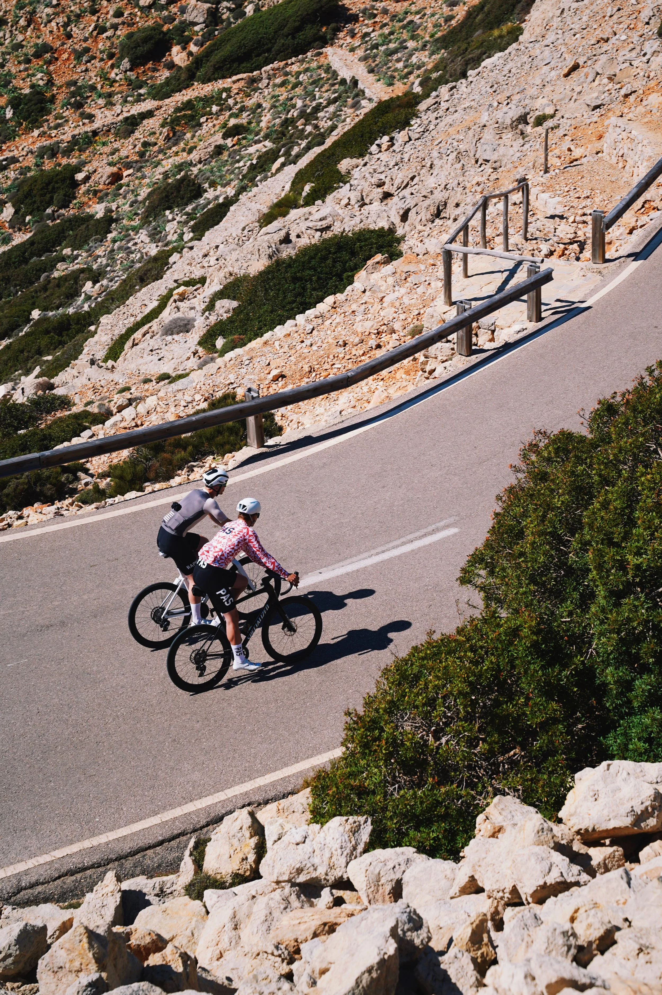 Two cyclists wearing helmets ride mountain bikes on a winding mountain road with rocky terrain and green bushes.