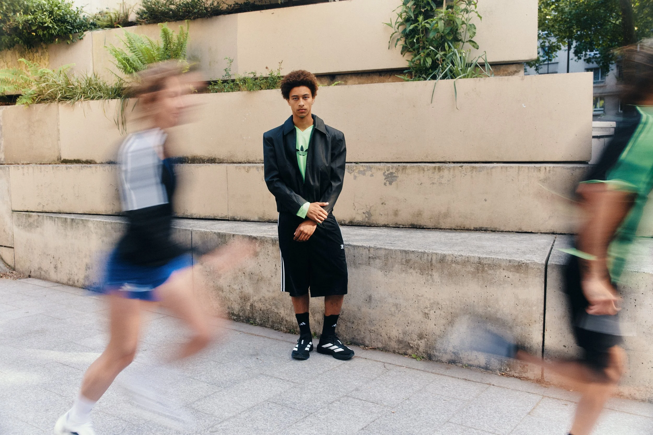 A young man standing against a concrete wall with greenery, while two blurred runners pass by him on the sidewalk.