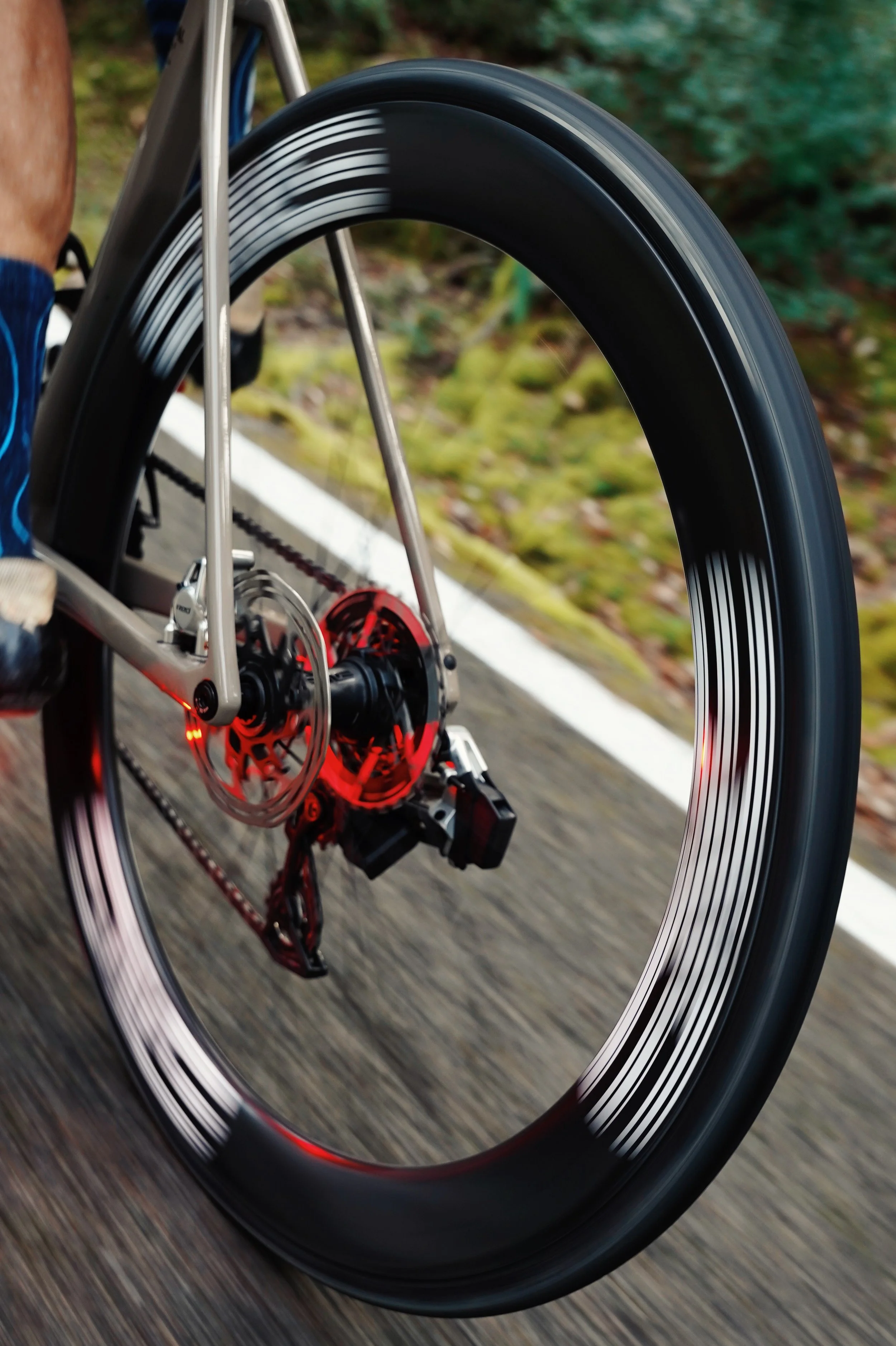 Close-up of a bicycle wheel with black and white striped pattern, showing motion blur as the bike rides on a paved road with nature in the background.