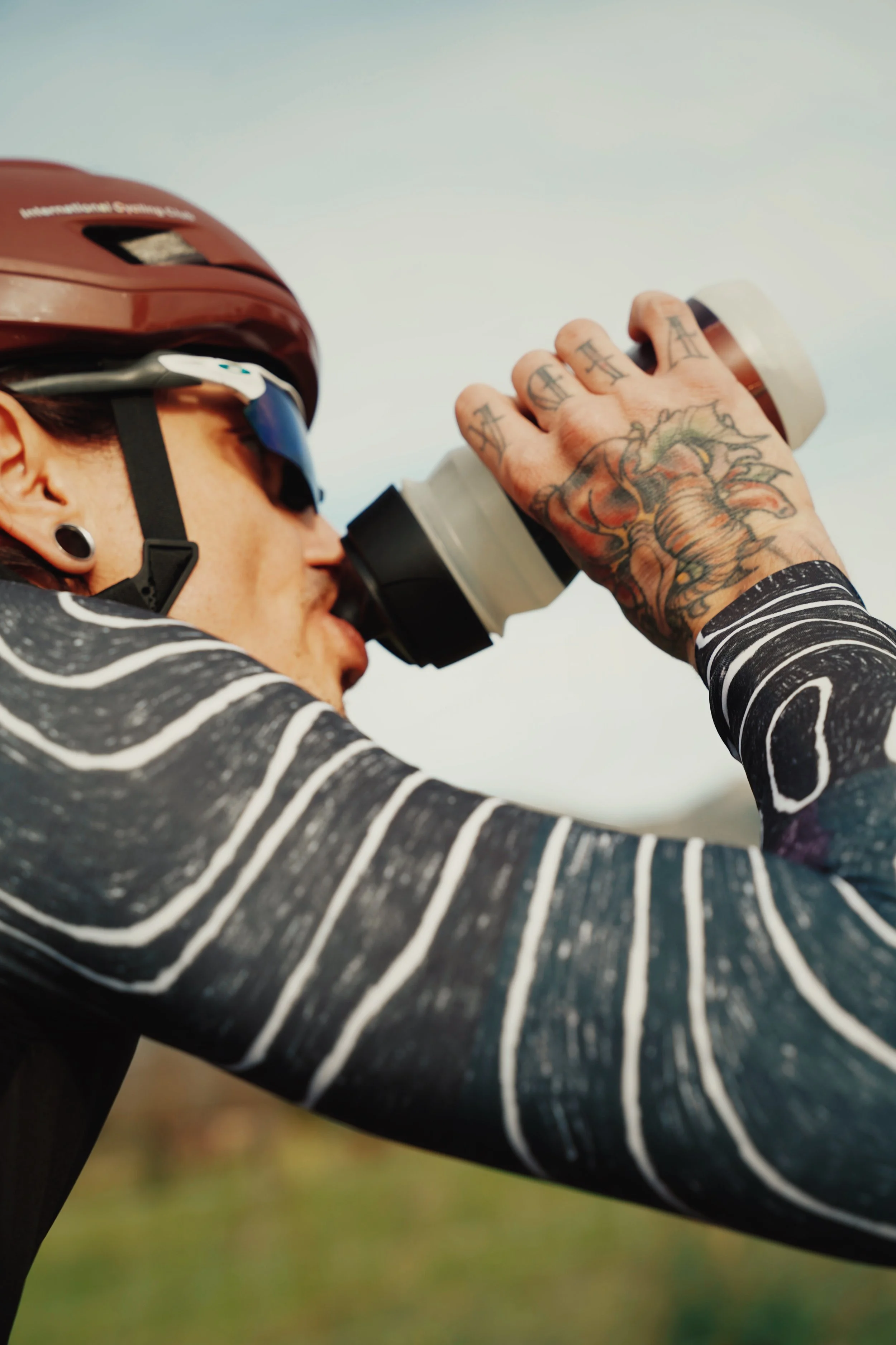 Person wearing a helmet and sunglasses drinking from a tumbler outdoors.