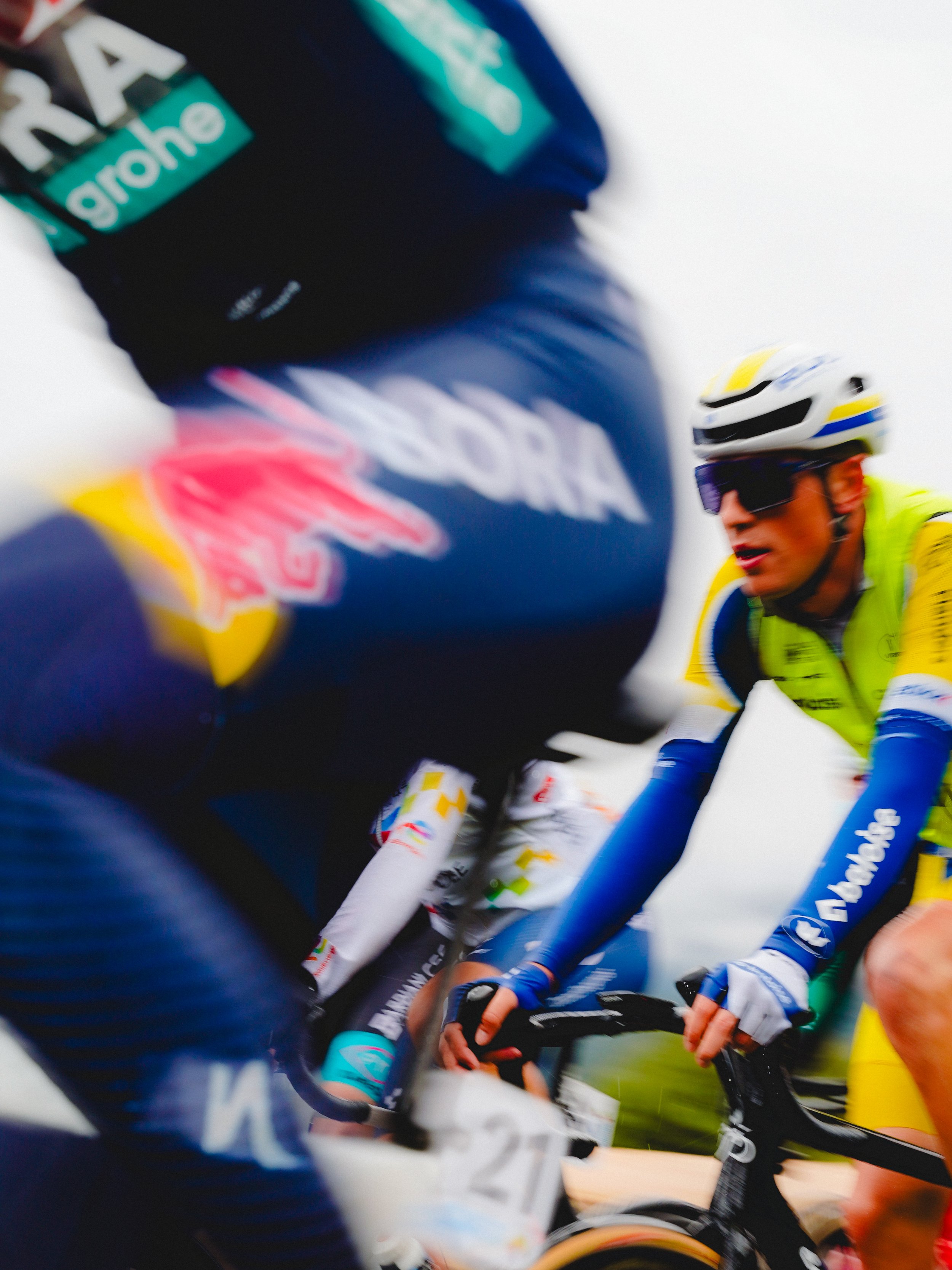 Close-up of professional cyclists racing, focusing on a cyclist in yellow and blue gear with a helmet and sunglasses, surrounded by other cyclists in colorful outfits.