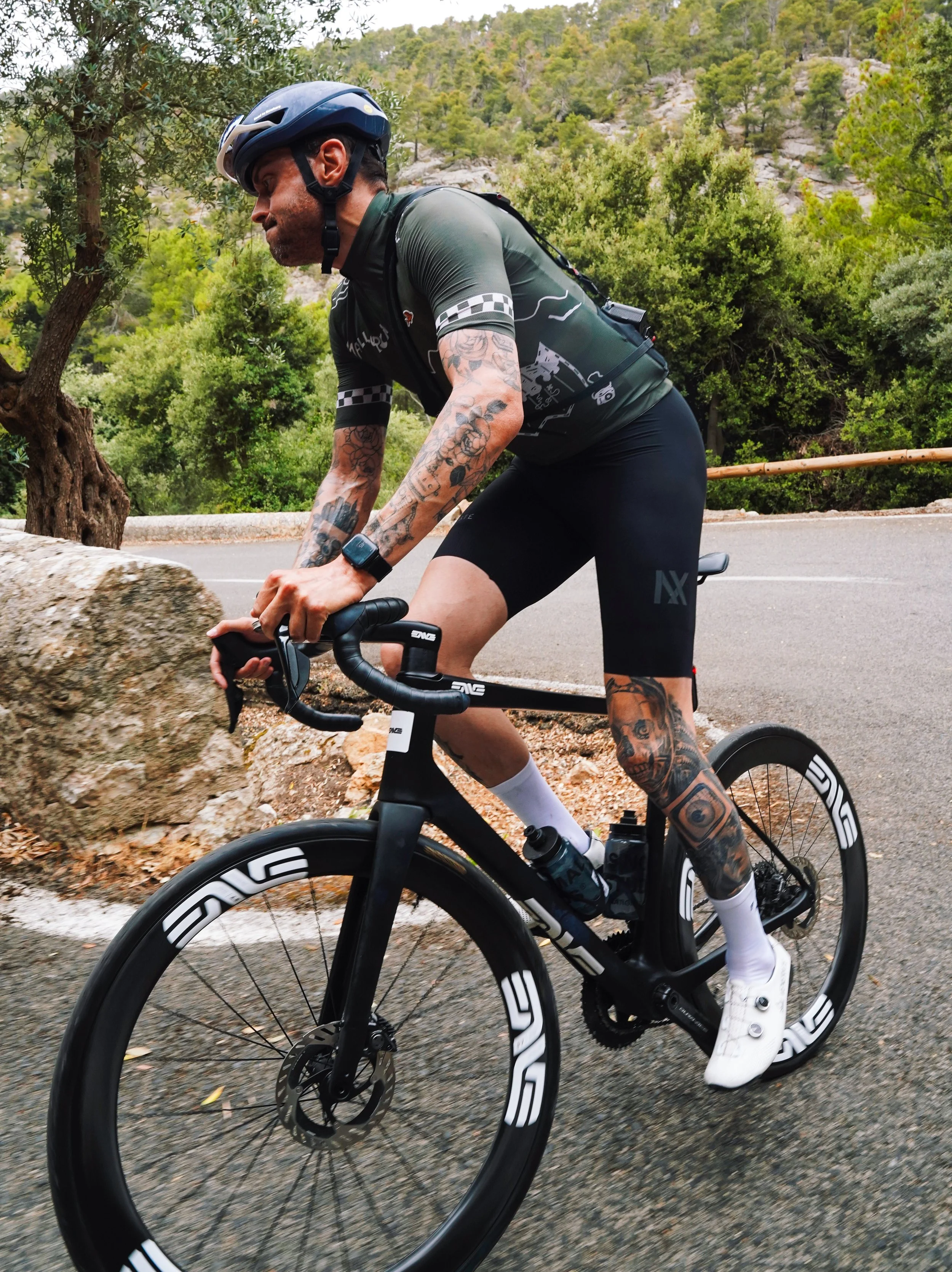 A man with tattoos wearing a helmet and cycling gear riding a black road bike with deep-section wheels on a mountain road surrounded by greenery.