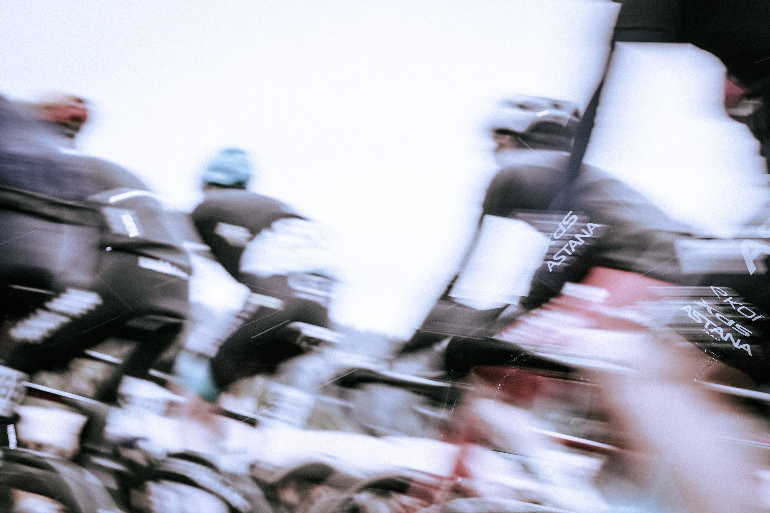 Blurred motion image of cyclists riding in a race, with some riders wearing black jerseys with white text, including 'ASTANA'.