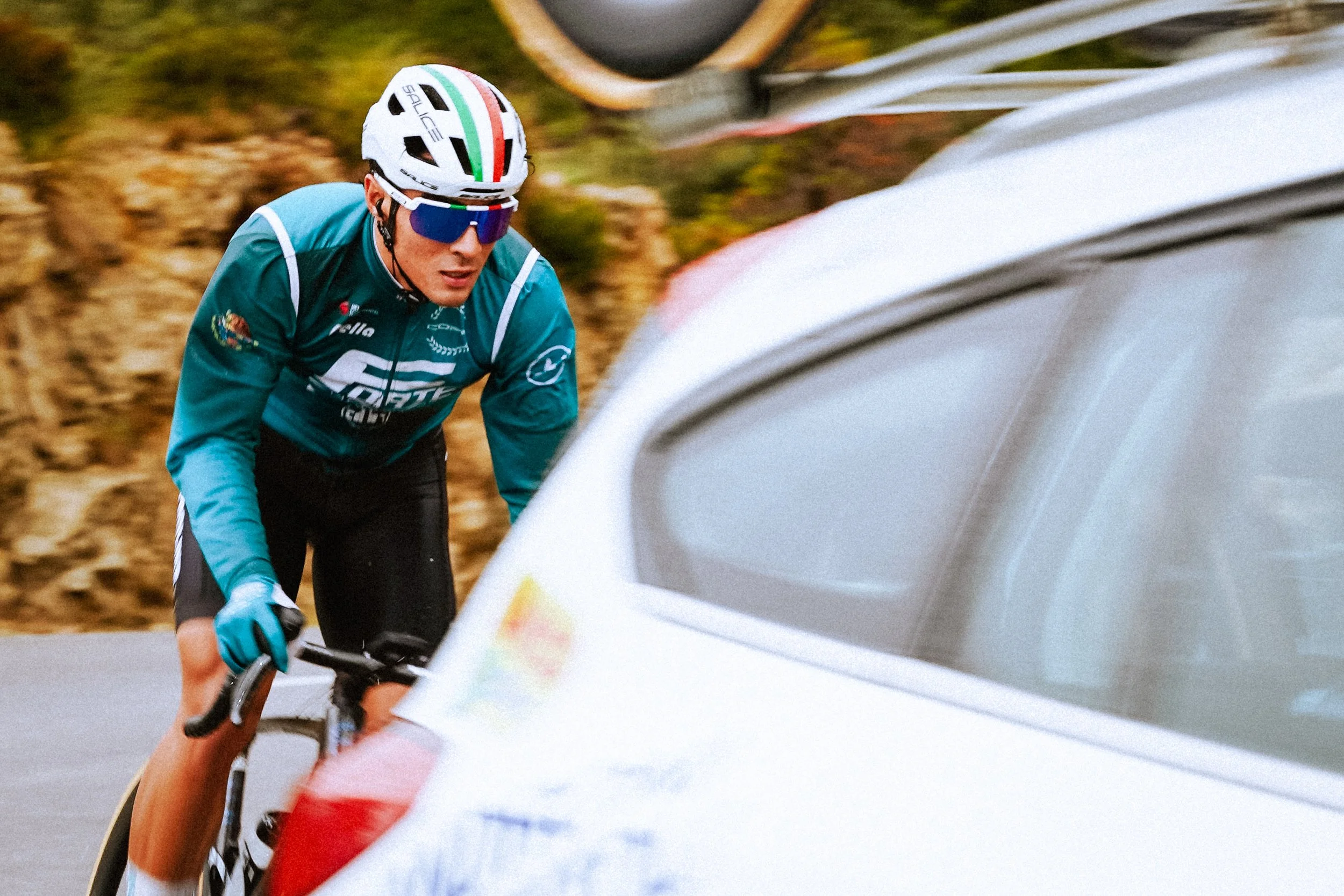 A professional cyclist wearing a teal jersey, sunglasses, and a helmet, riding his bike on a road with a white car partially in view and a blurred background of trees and dirt.