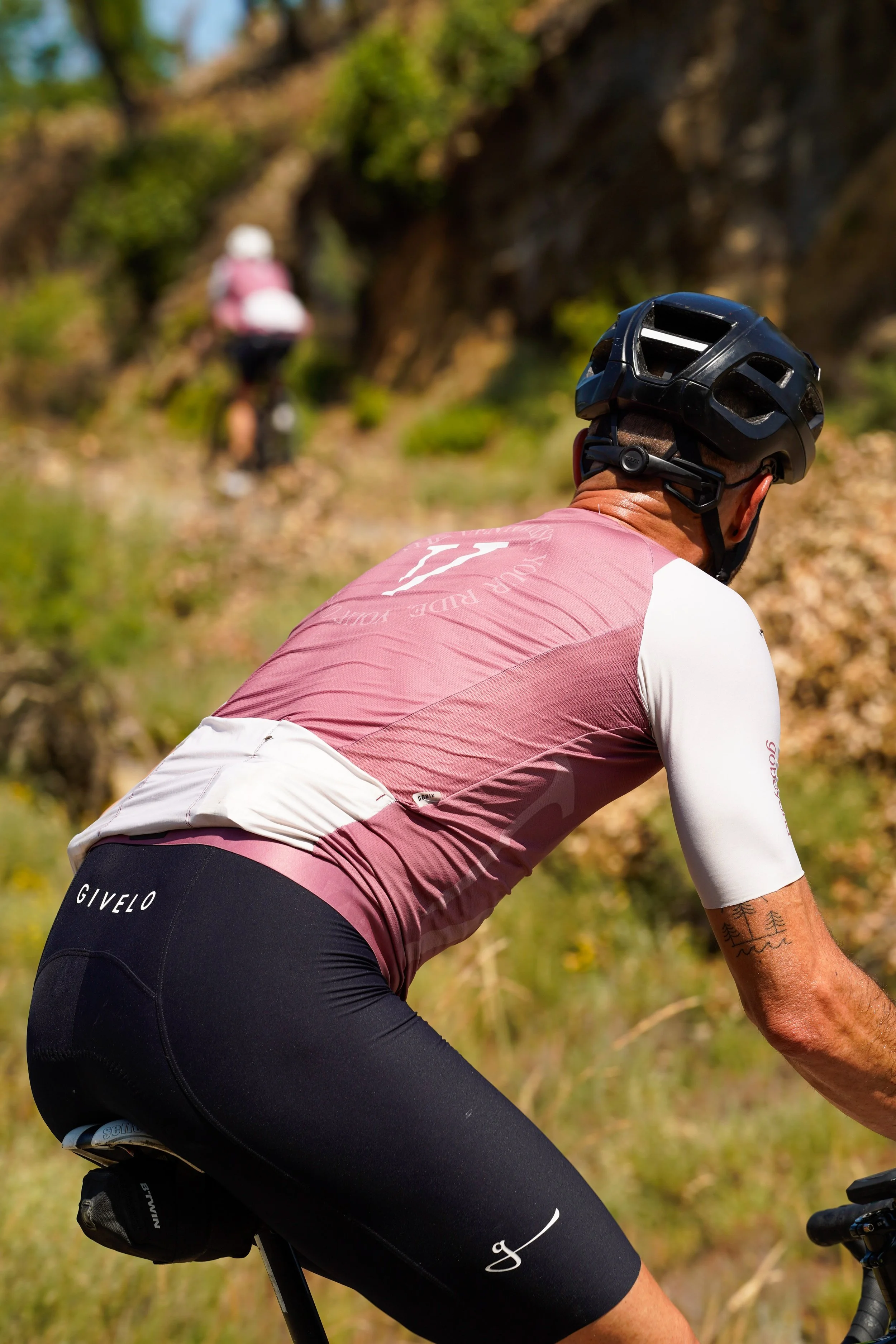 A cyclist wearing a black helmet, a pink and white cycling jersey, and black shorts riding on a dirt trail through a natural outdoor setting with green foliage and rocks. Another cyclist can be seen in the background.