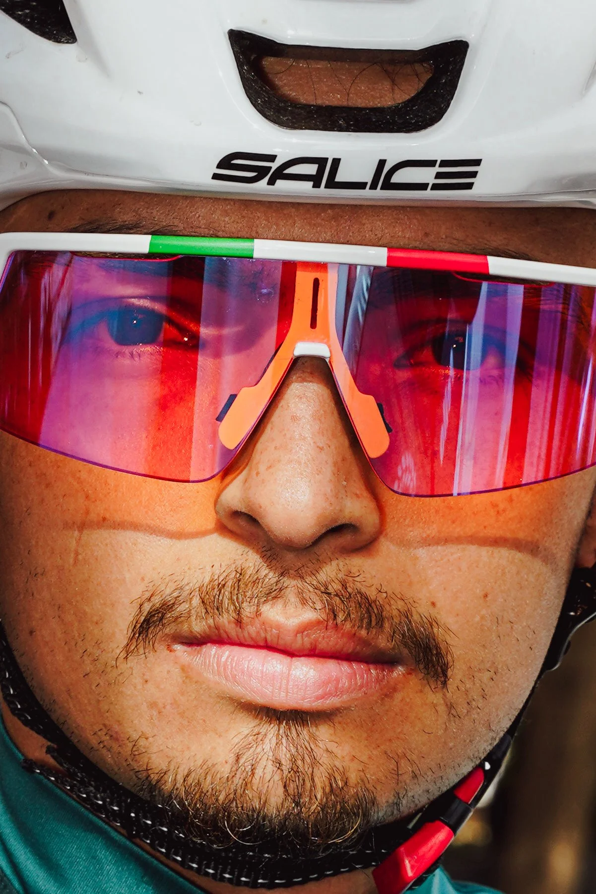 A close-up of a cyclist wearing an orange visor, reflective red sunglasses, a white helmet with black logo, and a teal shirt.