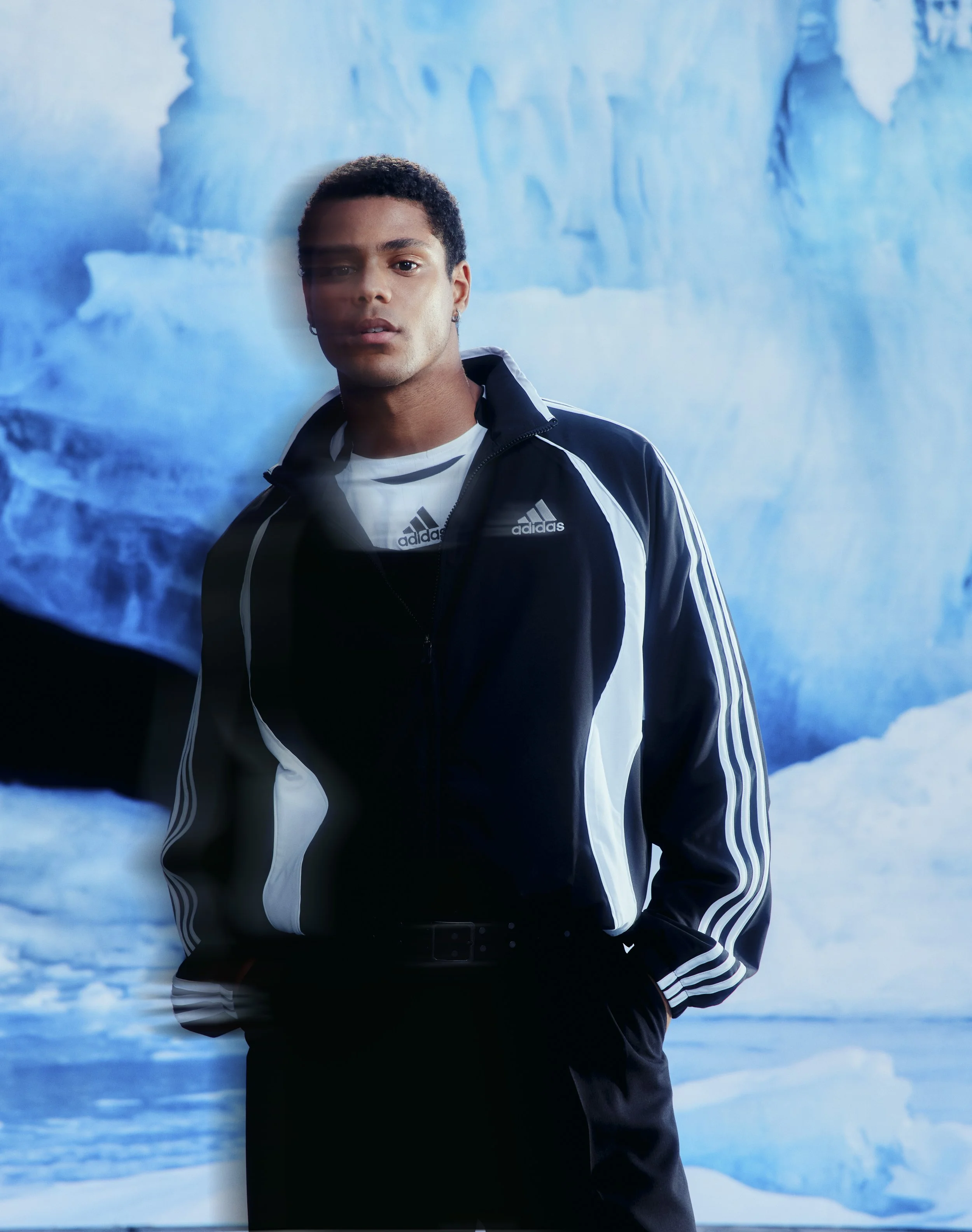 A young man with short curly hair and earrings stands in front of an icy blue glacier background. He is wearing a black and white Adidas track jacket over a white Adidas t-shirt.