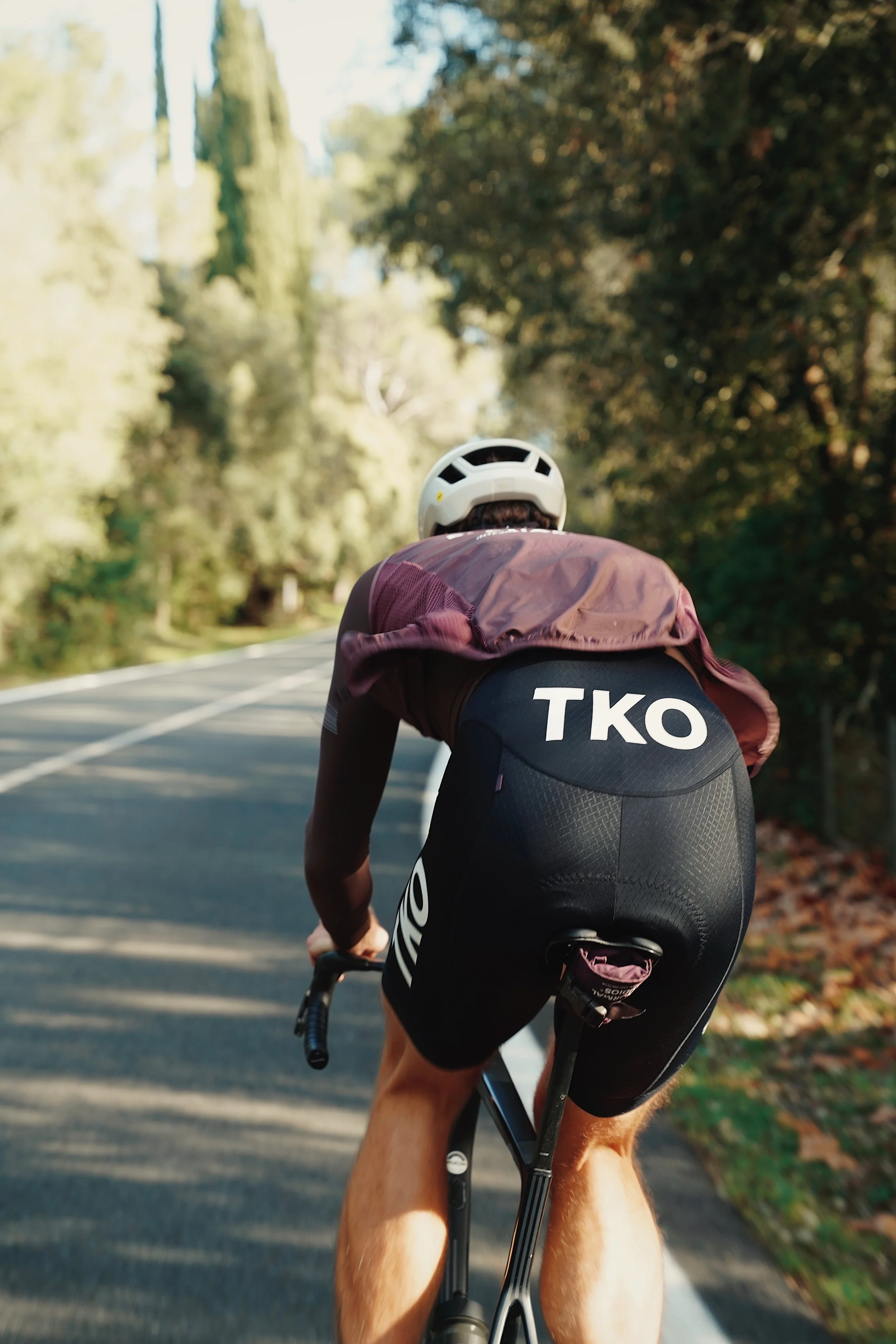 Cyclist riding on a rural road surrounded by trees, viewed from behind, wearing a helmet and black cycling gear with 'TKO' printed on the back.