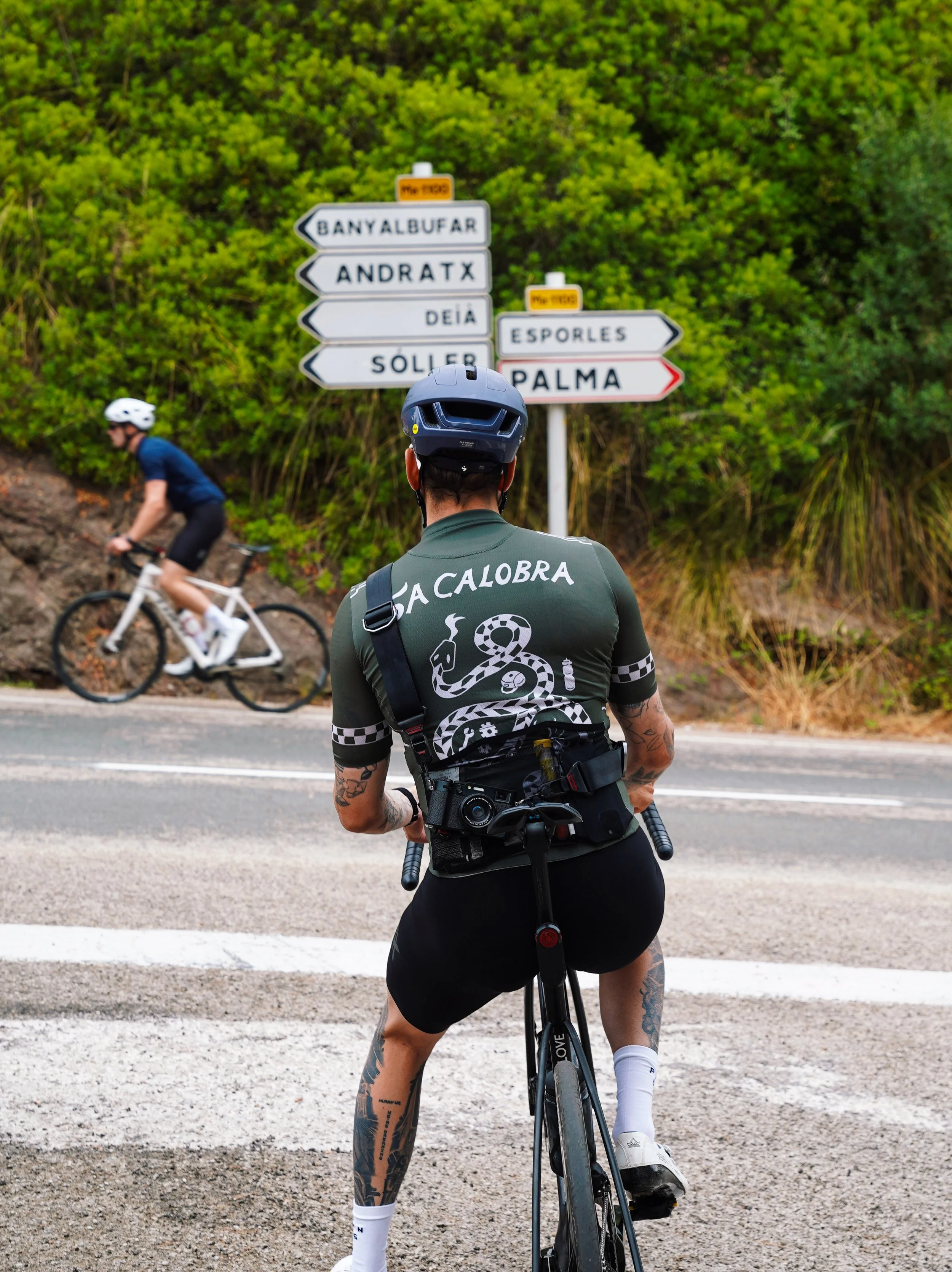 A cyclist wearing a dark helmet and a gray shirt with a snake graphic and the words 'La Calobra' is stopped at a pedestrian crosswalk, looking at a road sign with multiple directional signs. In the background, another cyclist is riding past, and ther