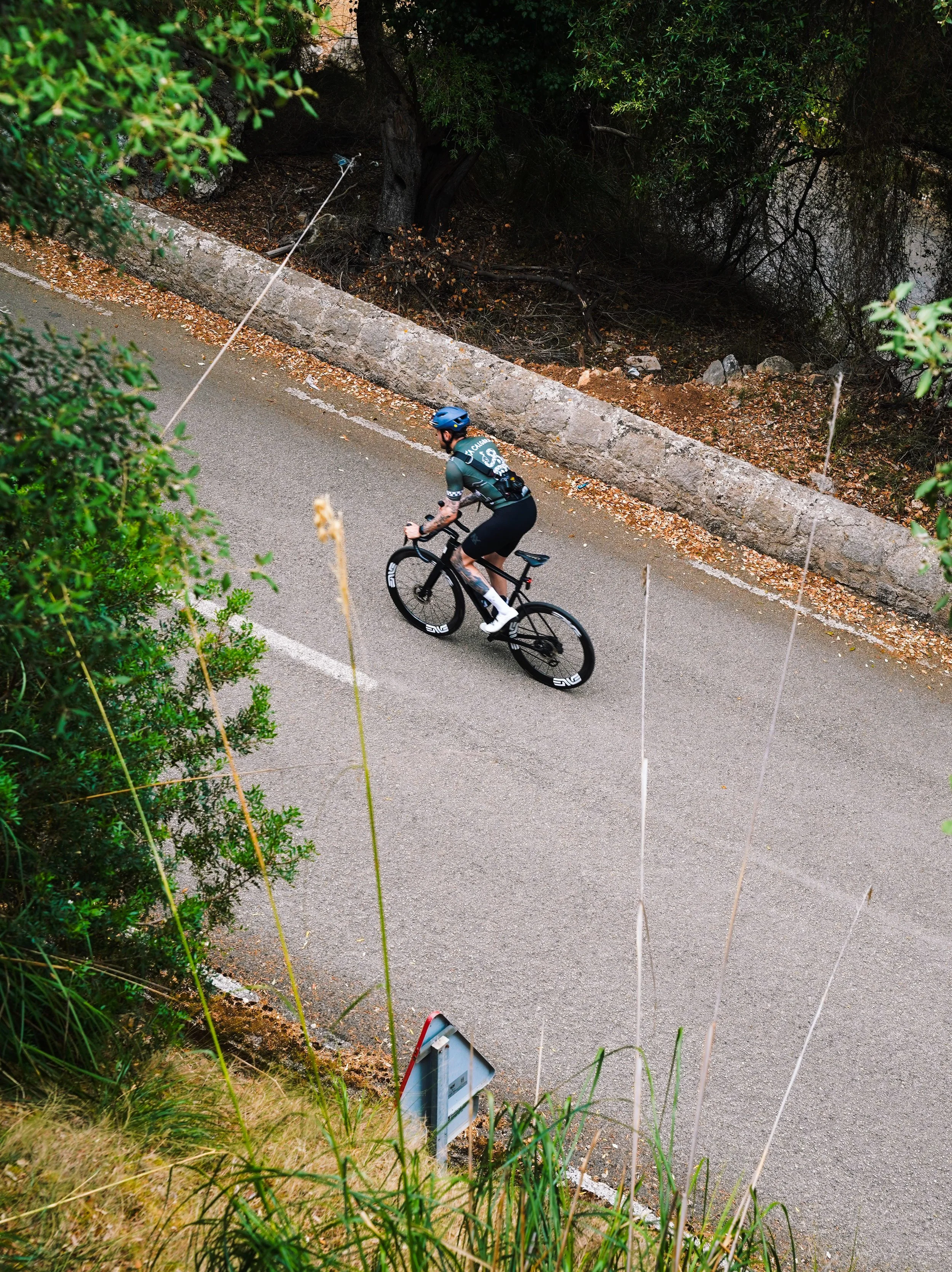 A cyclist wearing a black helmet and cycling attire riding a black bicycle on a paved road surrounded by greenery and trees.