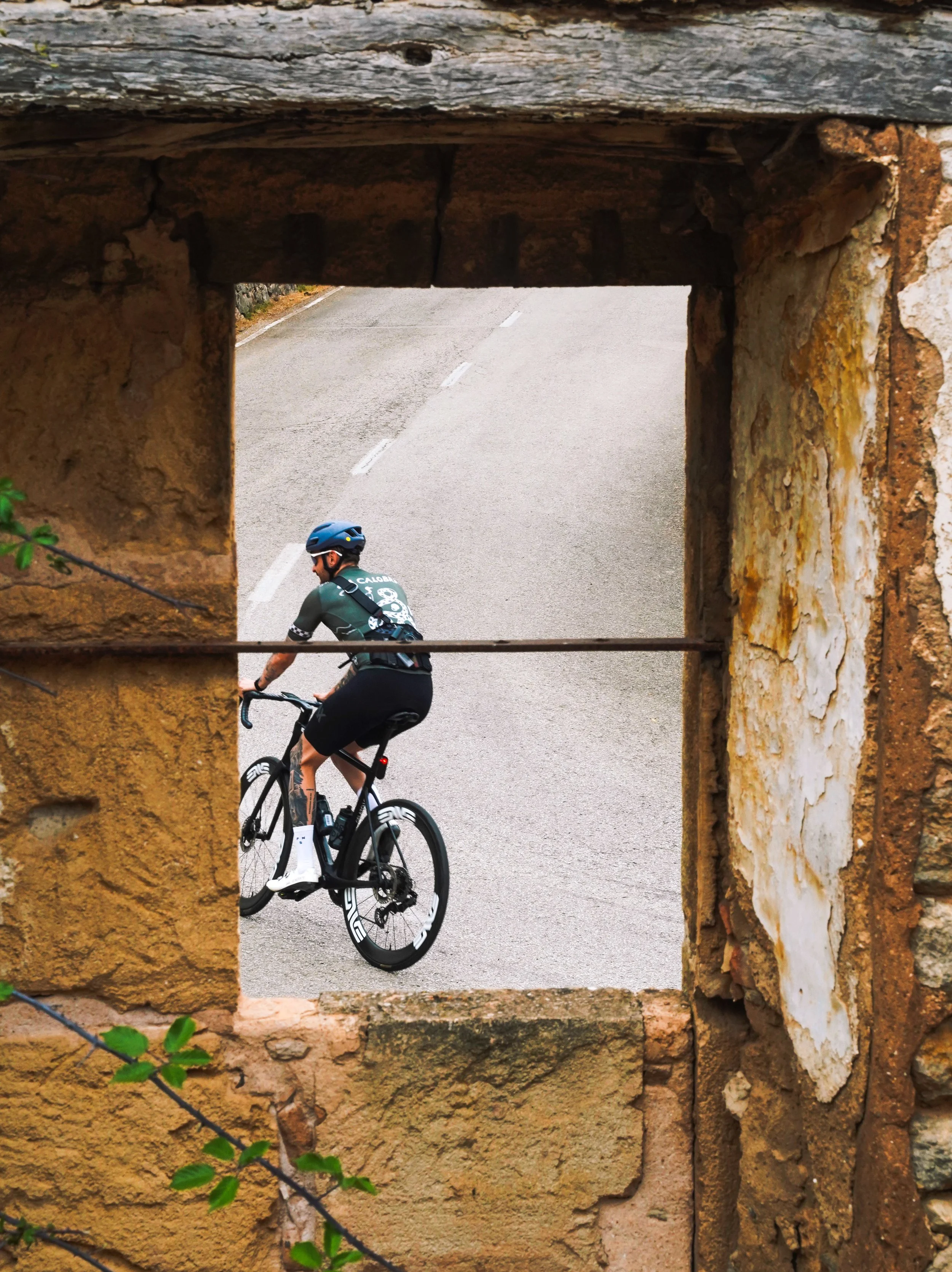 A cyclist riding a black bike on a paved road, viewed through a stone window with wooden trim, with some green leaves in the foreground.