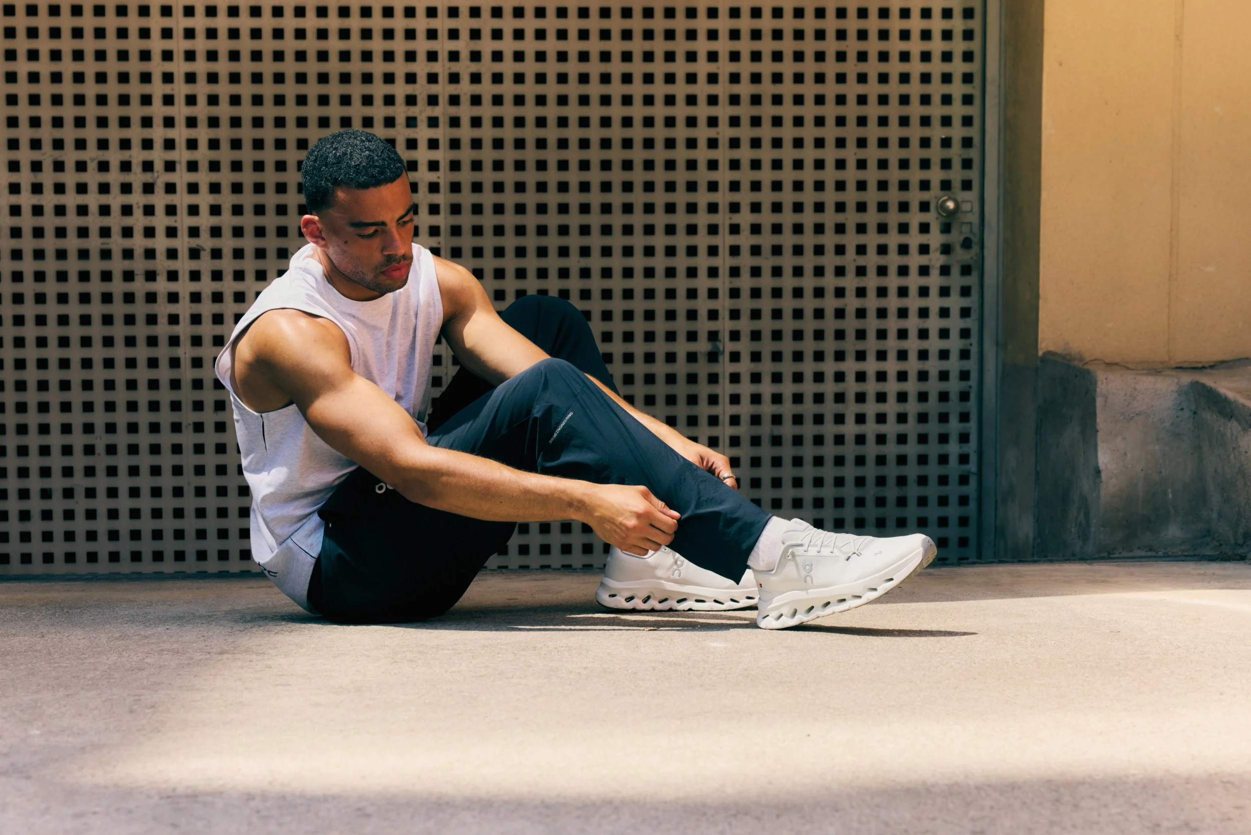 Man sitting on the ground tying his white sneakers, wearing a sleeveless shirt and black pants, against a textured wall background.