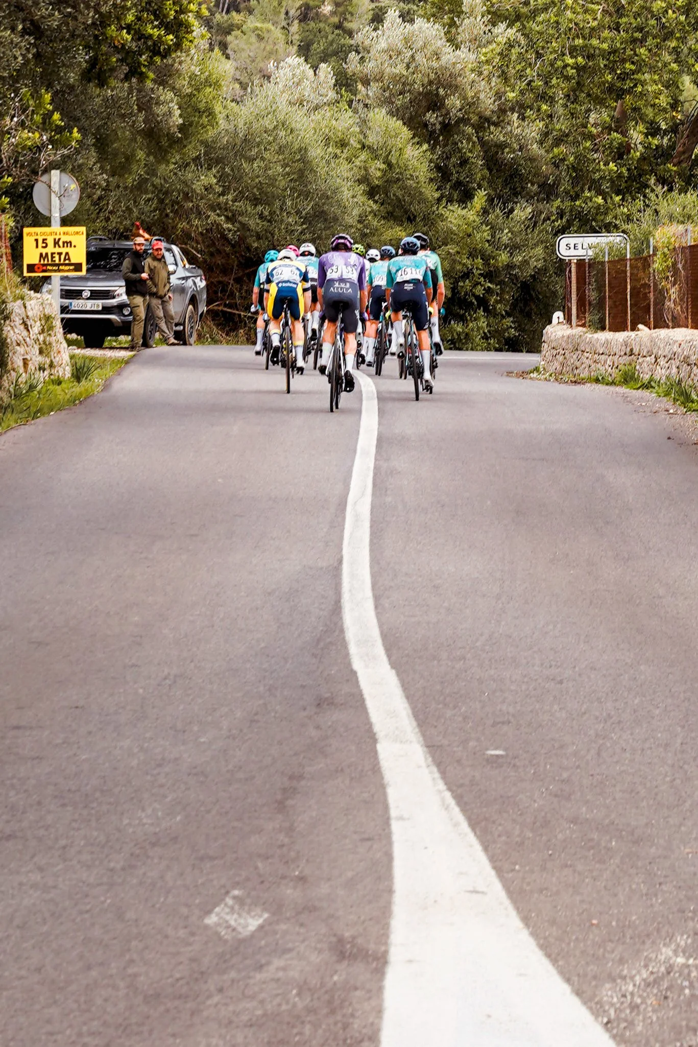 Group of cyclists riding on a rural road with trees on both sides, with two men standing next to a parked car on the left and a street sign that reads 'SE' on the right.