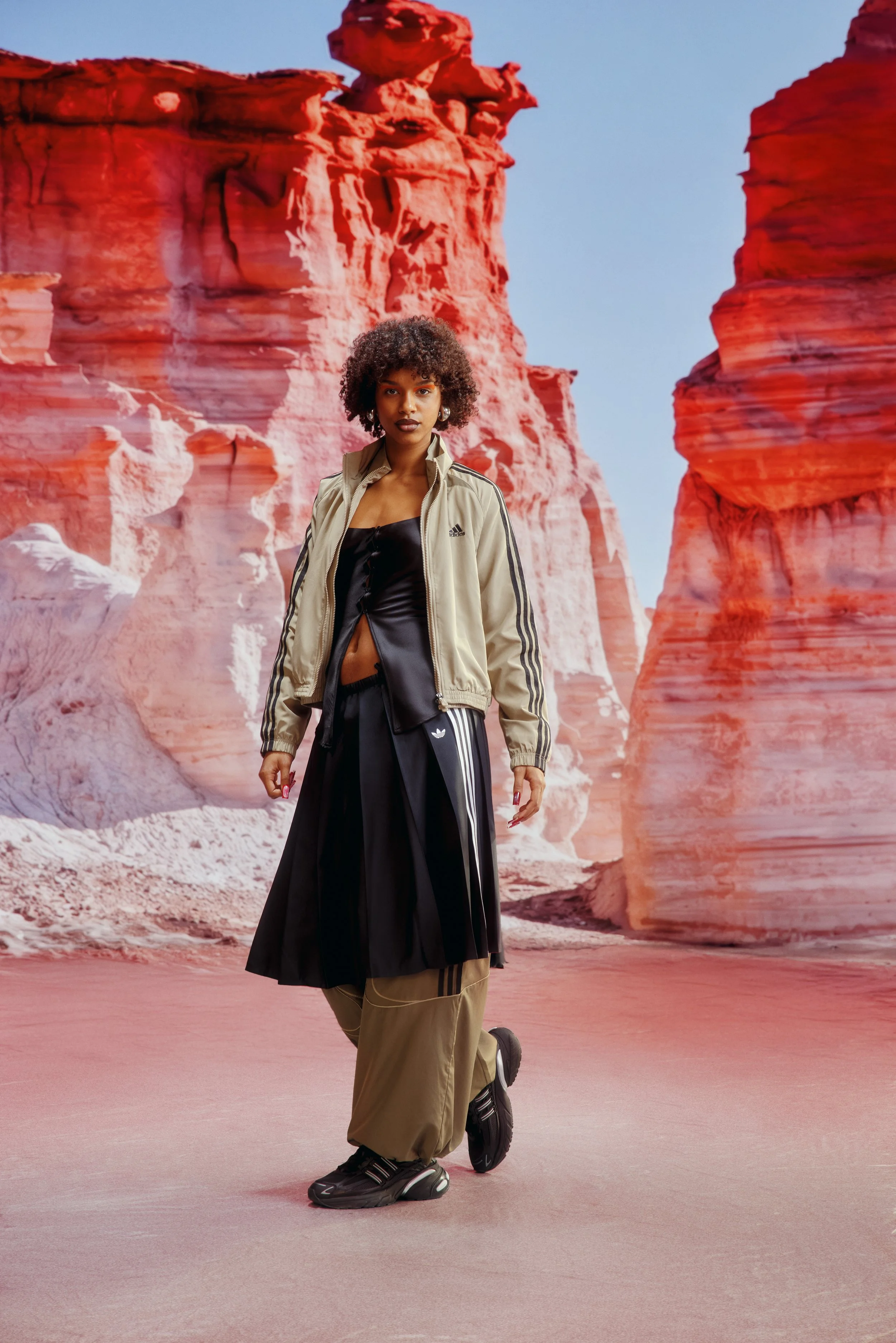 A woman standing outdoors in front of red rock formations, dressed in athletic clothing, including a beige Adidas jacket, black midi skirt, and sneakers.