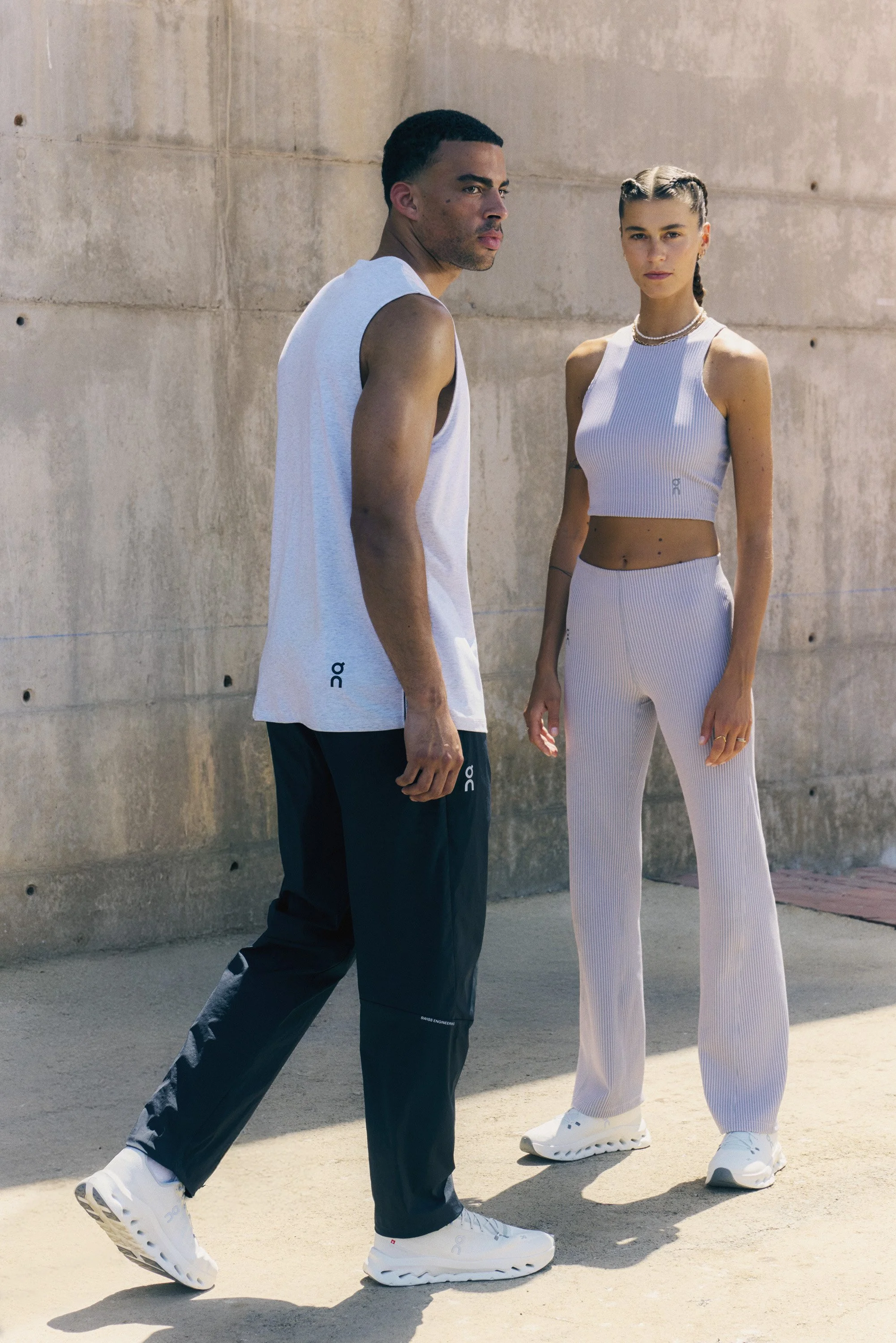 Two people dressed in athletic wear standing outside near a concrete wall. The man is looking off to the side, and the woman is looking forward.