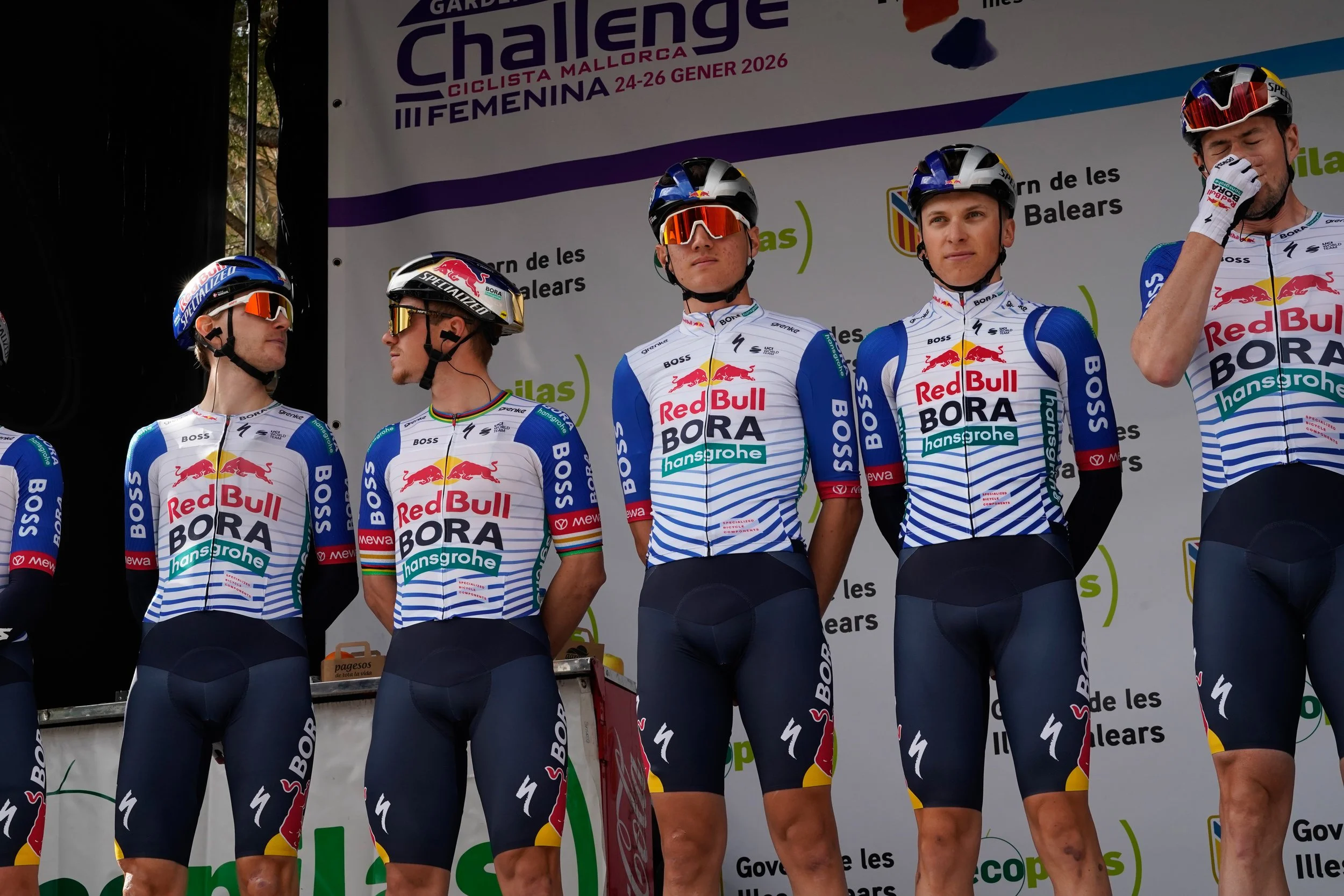 Cyclists in team uniforms standing on stage during a ceremony at a cycling event, with a backdrop displaying event details, including dates and location, and sponsor logos.