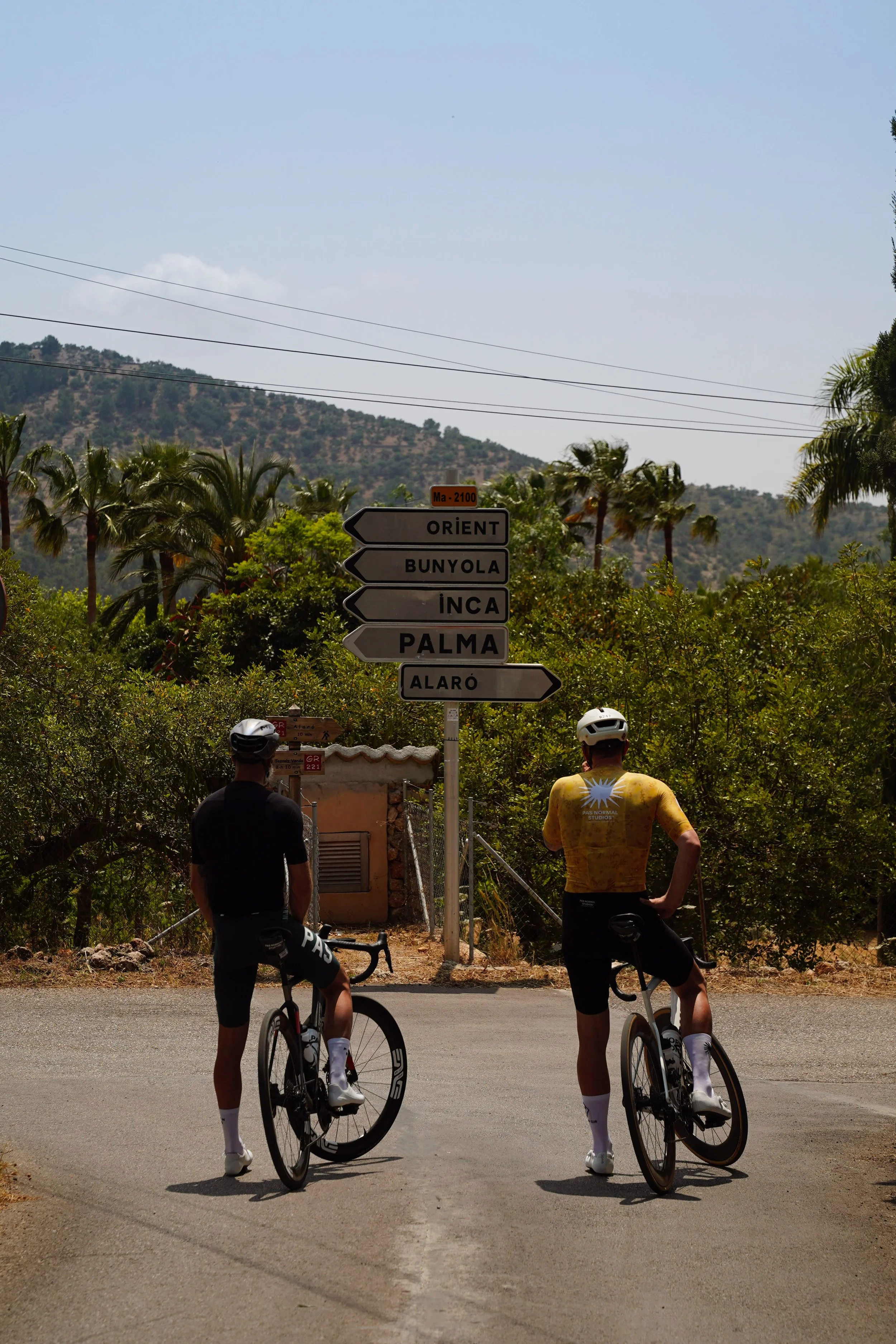 Two cyclists with helmets and cycling gear stand on a road, looking at a directional signpost showing directions to Oriént, Bunyola, Inca, Palma, and Alaró, in a rural area with trees and mountains in the background.
