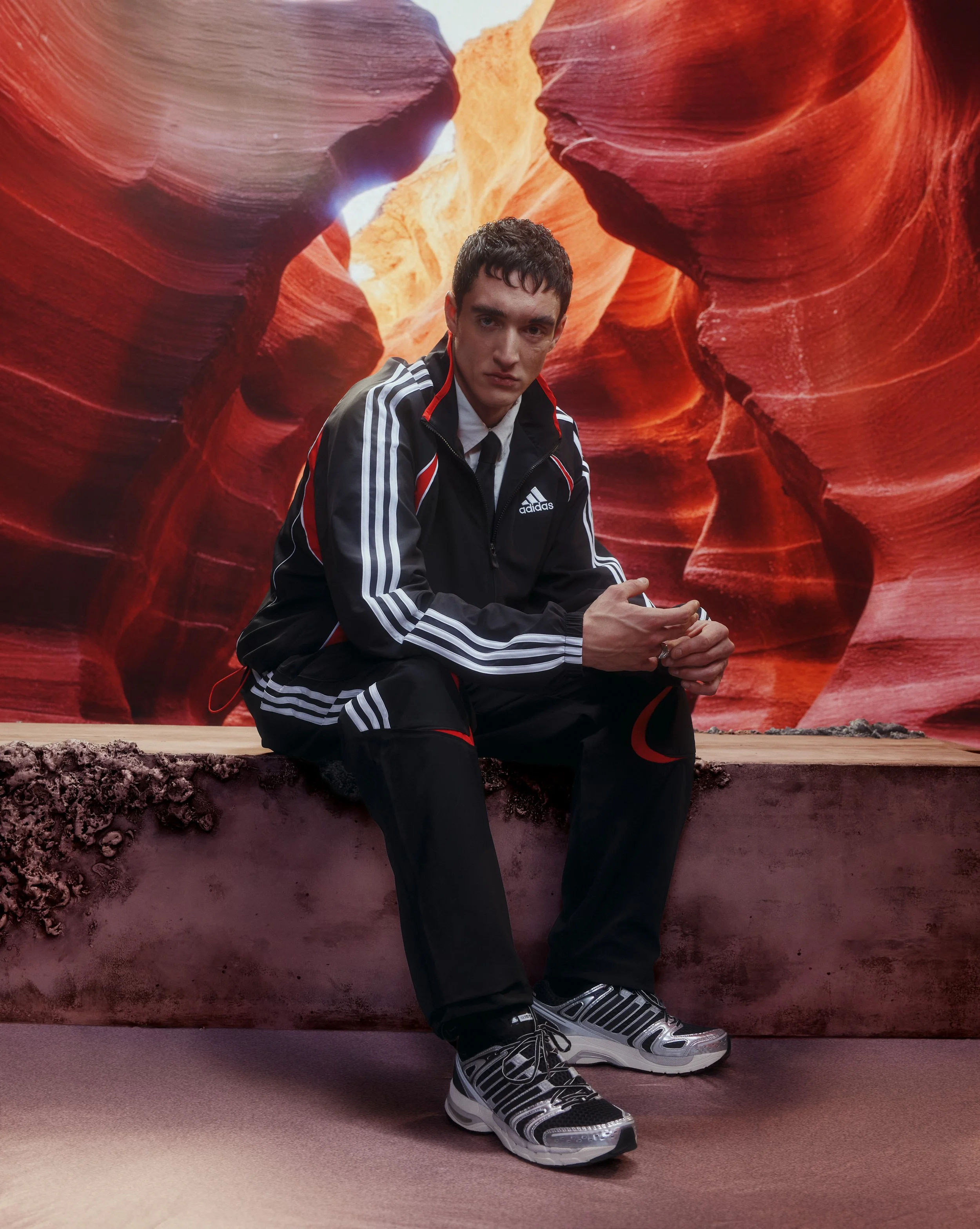 A young man in a black Adidas tracksuit with white and red accents, sitting on a concrete ledge in front of a backdrop resembling the Antelope Canyon with orange and red rock formations.