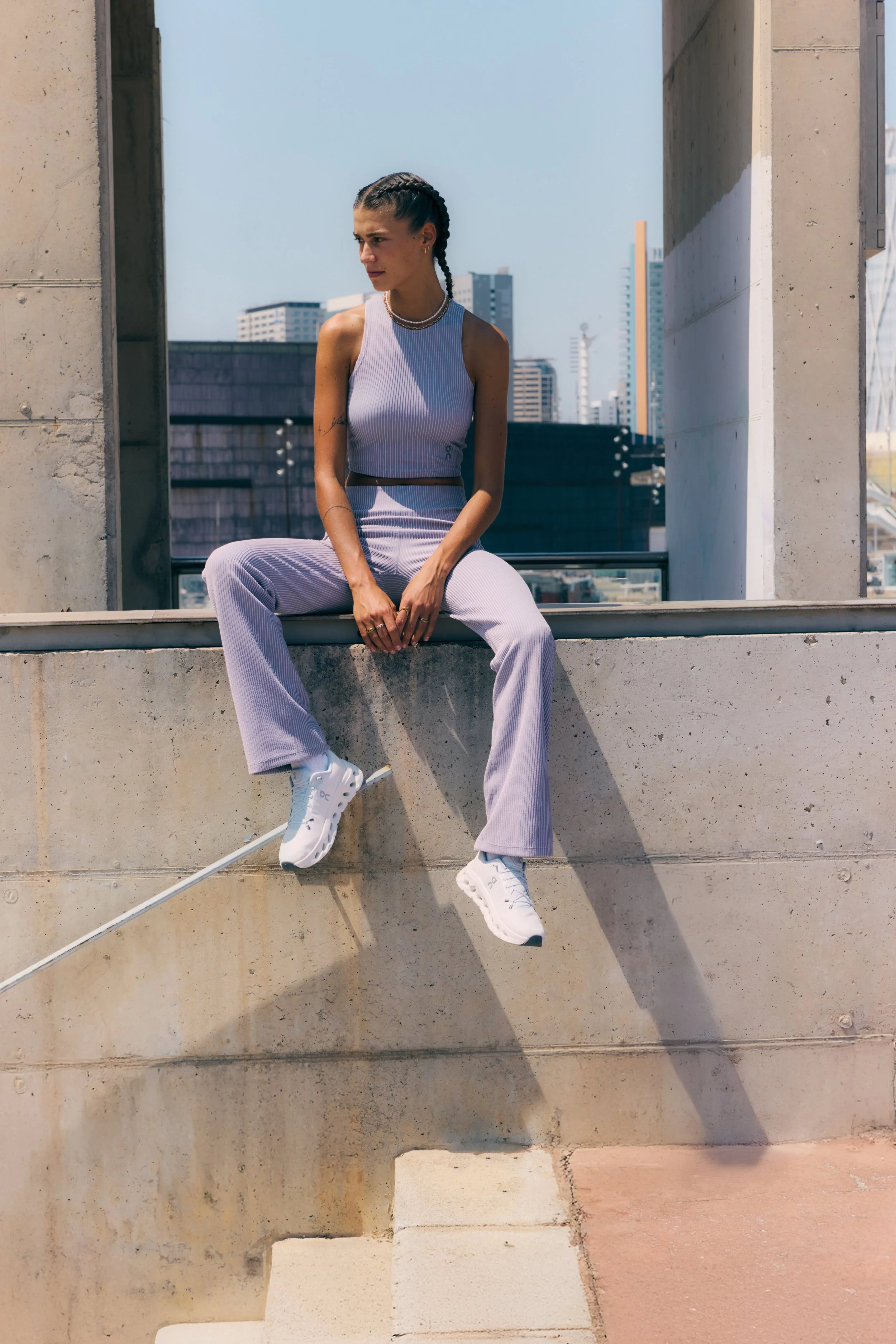 A woman wearing a sleeveless, purple, ribbed workout outfit and white sneakers sitting on a concrete ledge in an urban setting with tall buildings in the background.
