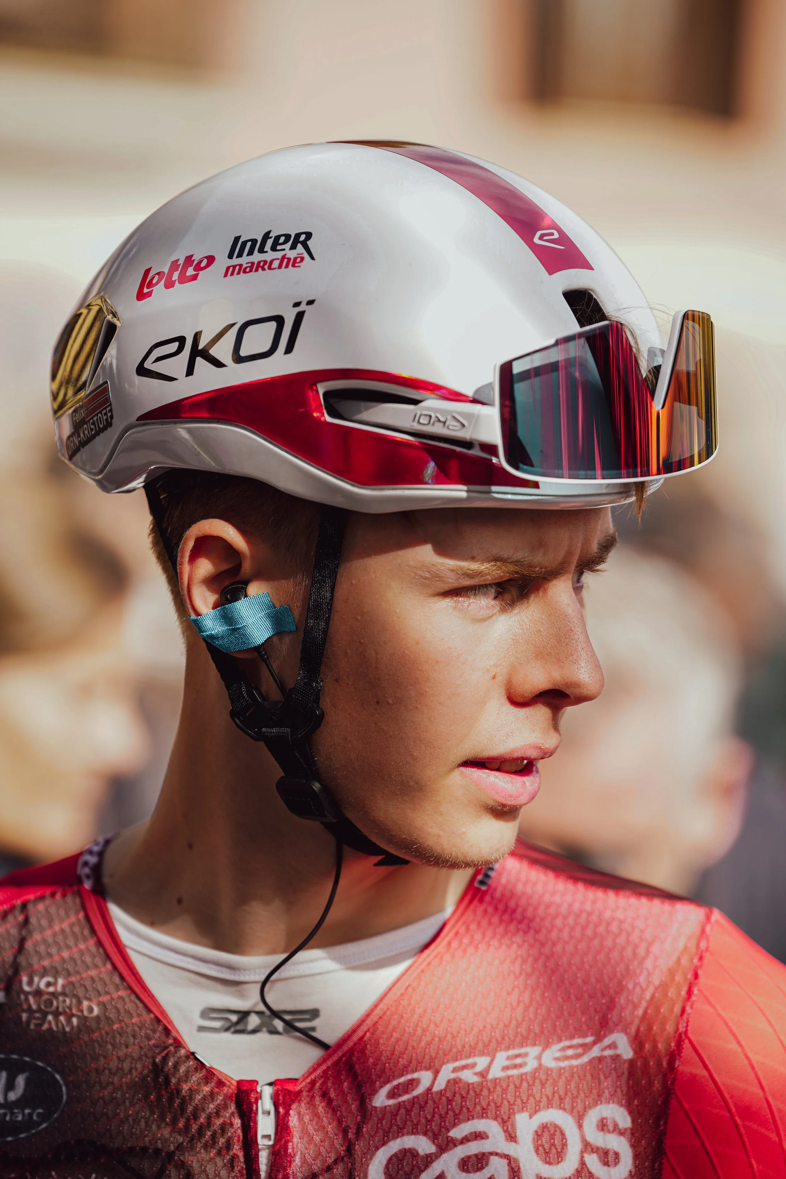 Close-up of a cyclist wearing a white helmet with red accents, orange tinted sunglasses, and a red cycling jersey, looking to the side with a serious expression.